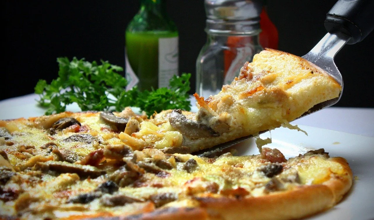 Slice of mushroom and cheese pizza being lifted with a pizza cutter on a white plate, with green herbs and condiments in the background.