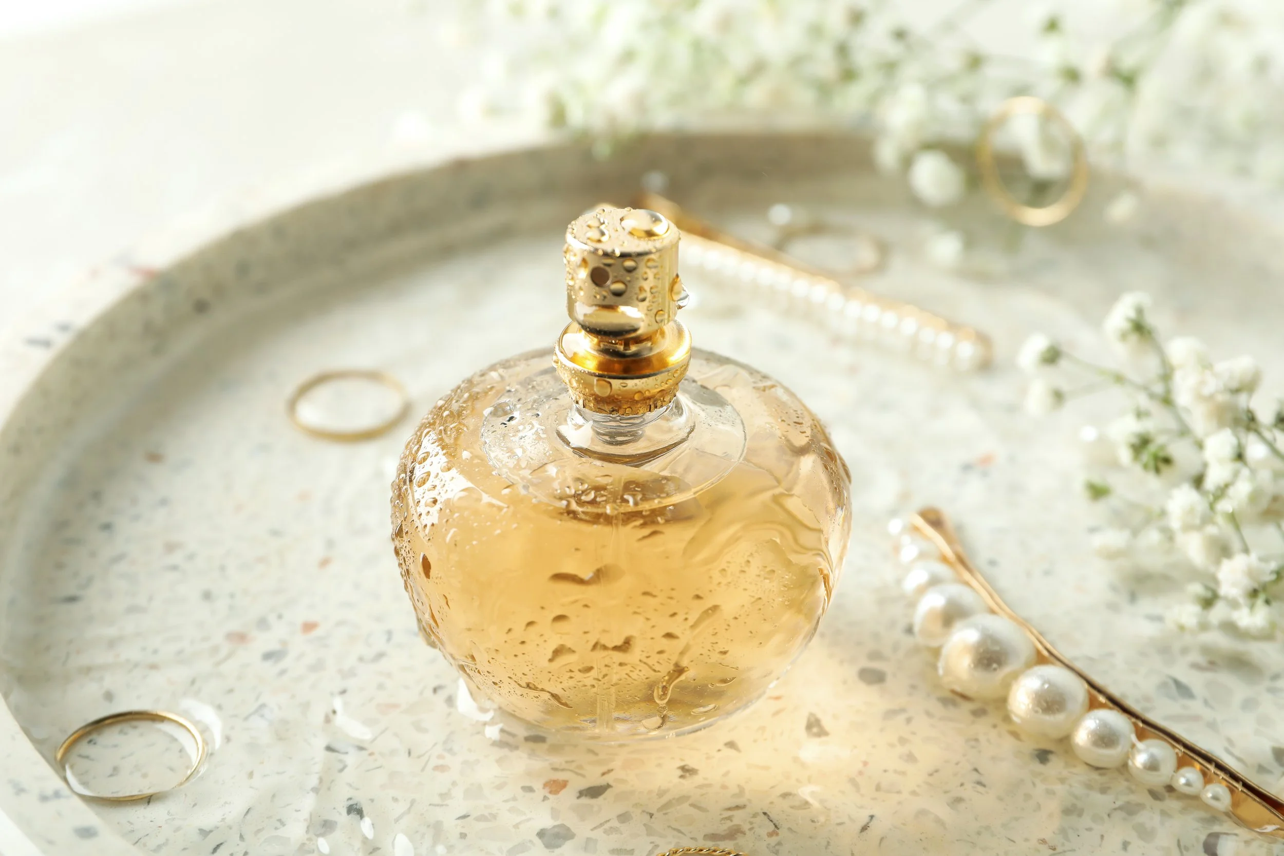 A perfume bottle with a golden cap and liquid inside, surrounded by pearl jewelry and white flowers, placed on a silver tray with water droplets on the bottle.