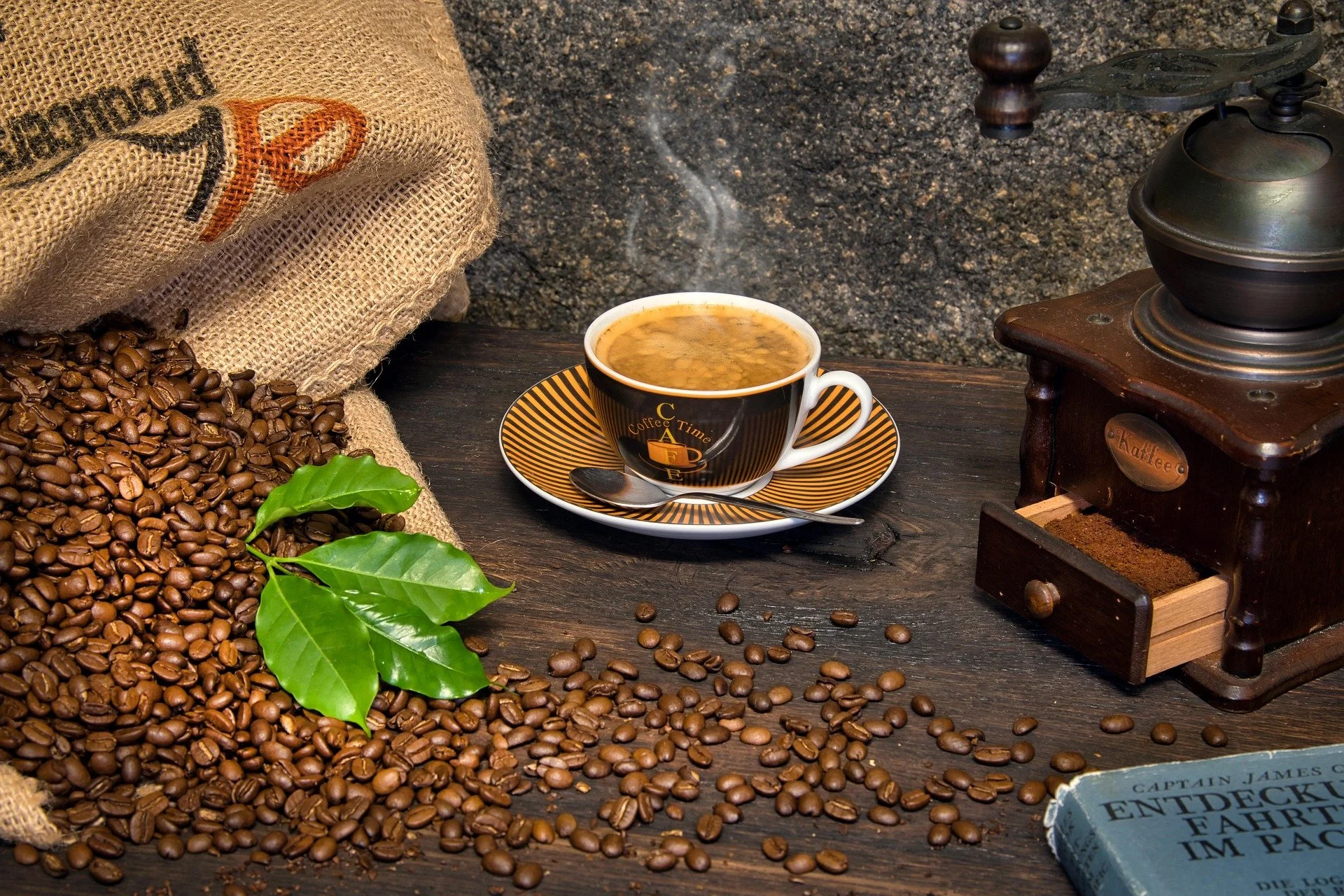 A steaming cup of coffee in a black cup with a saucer, placed on a dark wooden table along with scattered coffee beans, a vintage coffee grinder, a burlap sack of coffee beans, and a green coffee plant leaf.