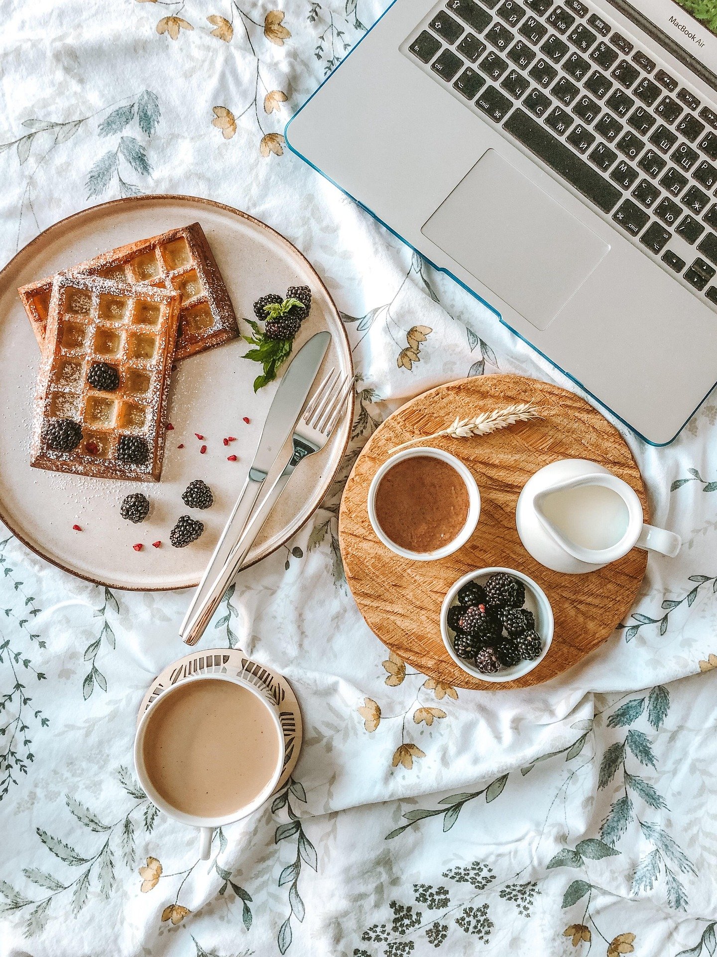 Breakfast setup with waffles topped with blackberries on a white plate, two cups of coffee, a small bowl of blackberries, a small jug of milk, a small container of chocolate spread, and a laptop on a floral bedsheet.