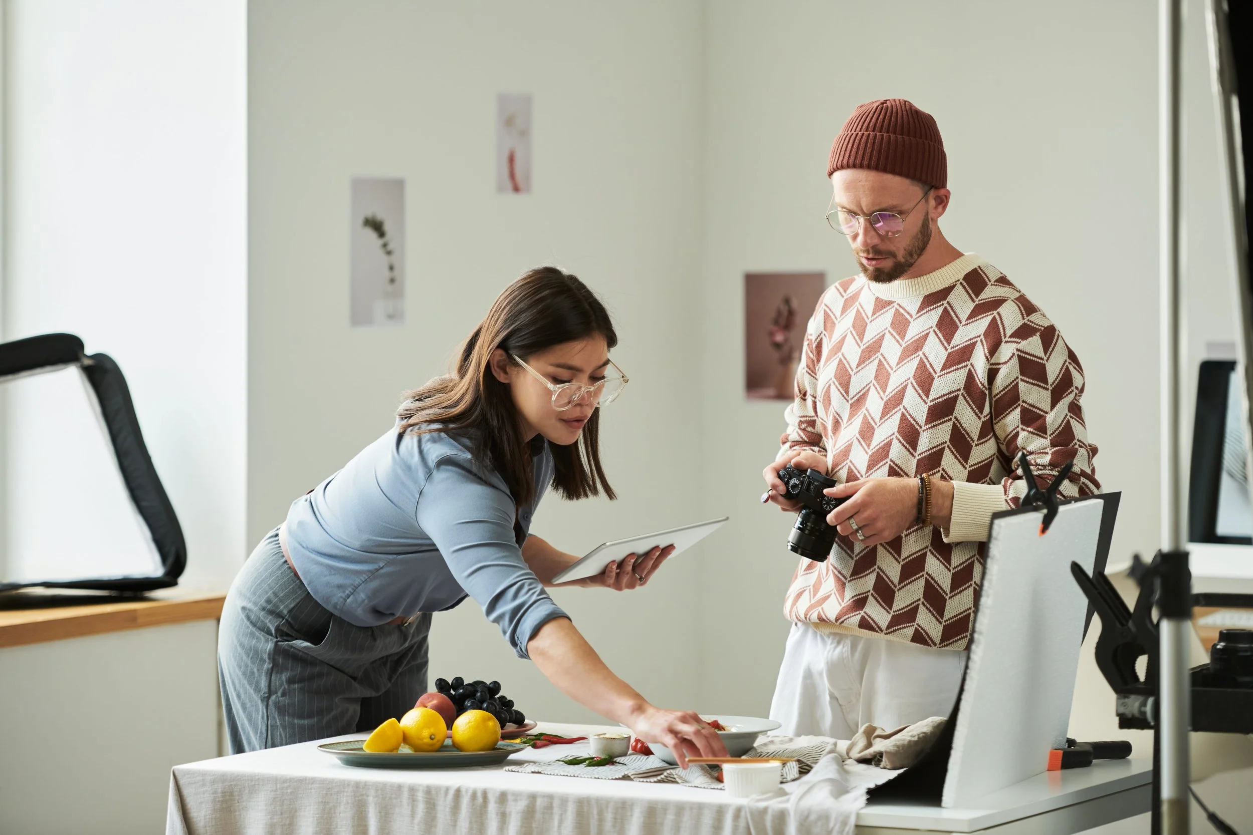 Two people, a woman and a man, working together in a studio or kitchen. The woman is reaching for fruit on a table, while the man holds a camera and watches. The table has lemons, grapes, and a small bowl. The woman is holding a tablet, and there is 