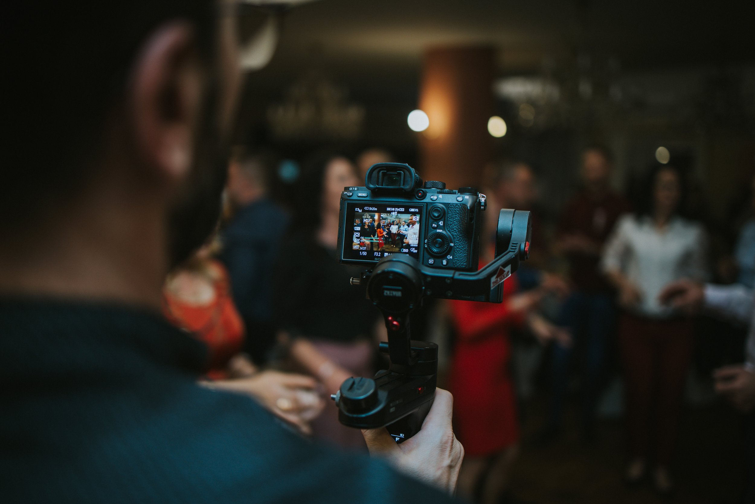 Photographer recording an indoor social event with a professional camera, with blurred group of people in the background.