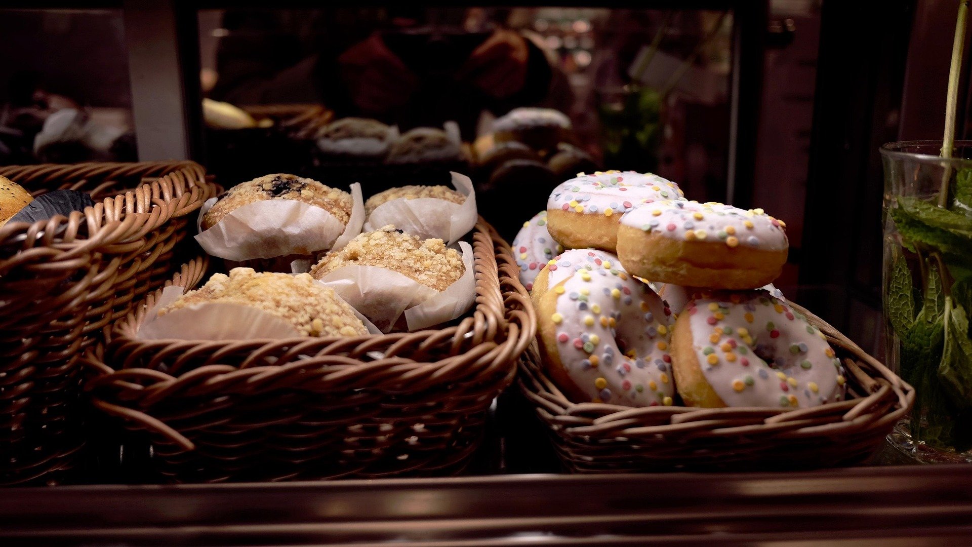 Wicker baskets containing muffins and donuts with colorful sprinkles, displayed behind a glass in a bakery or café setting.