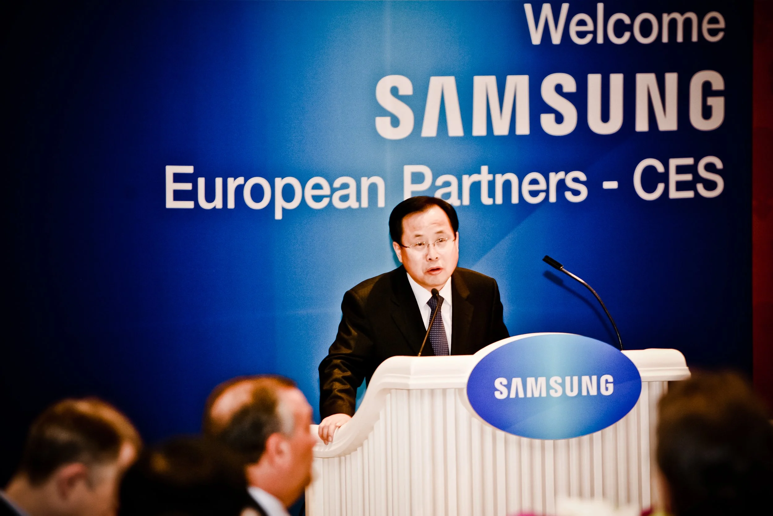 A man in a black suit and tie speaking at a podium with a blue Samsung logo. Behind him is a blue backdrop with white text that reads, "Welcome Samsung European Partners - CES." There are attendees in the foreground listening.