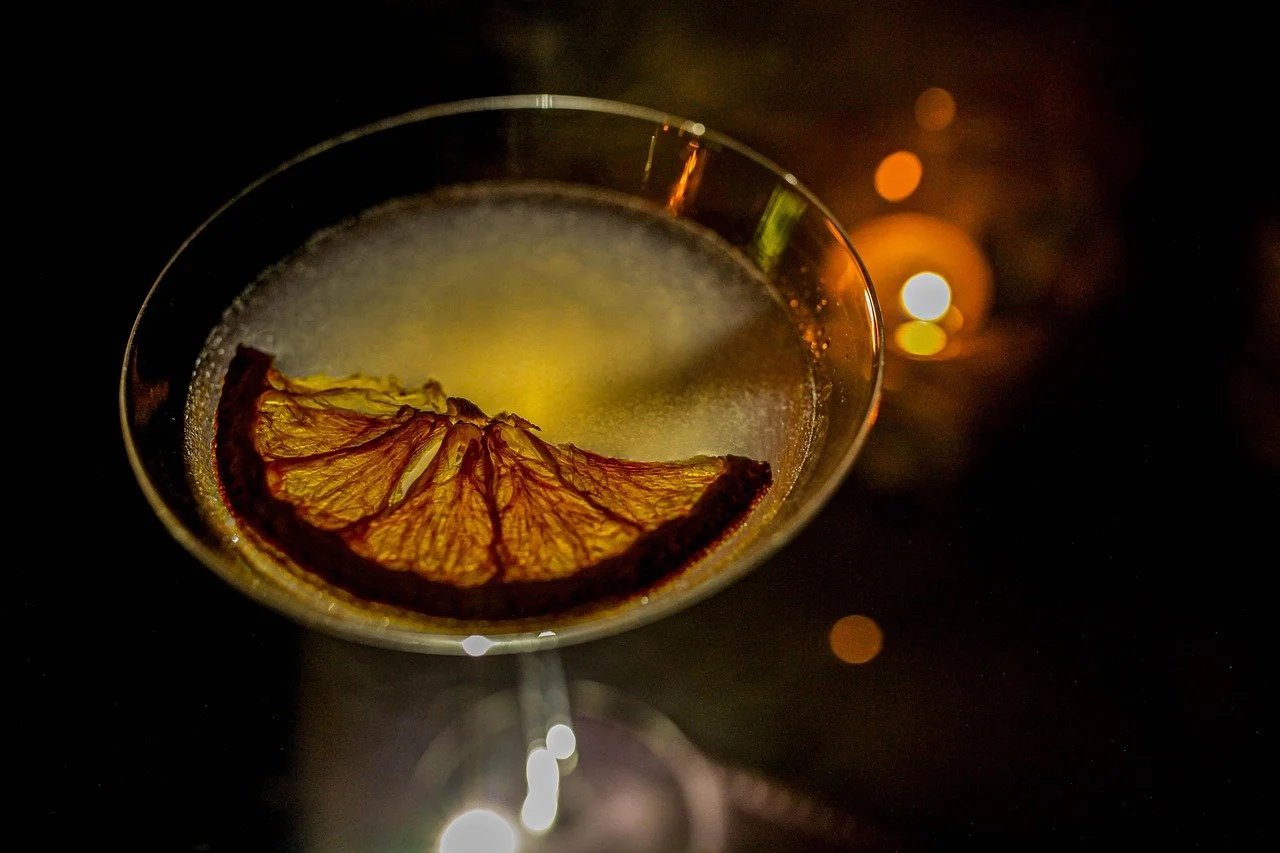 A cocktail glass with a slice of dried orange inside, illuminated in a dark setting with soft bokeh lights in the background.