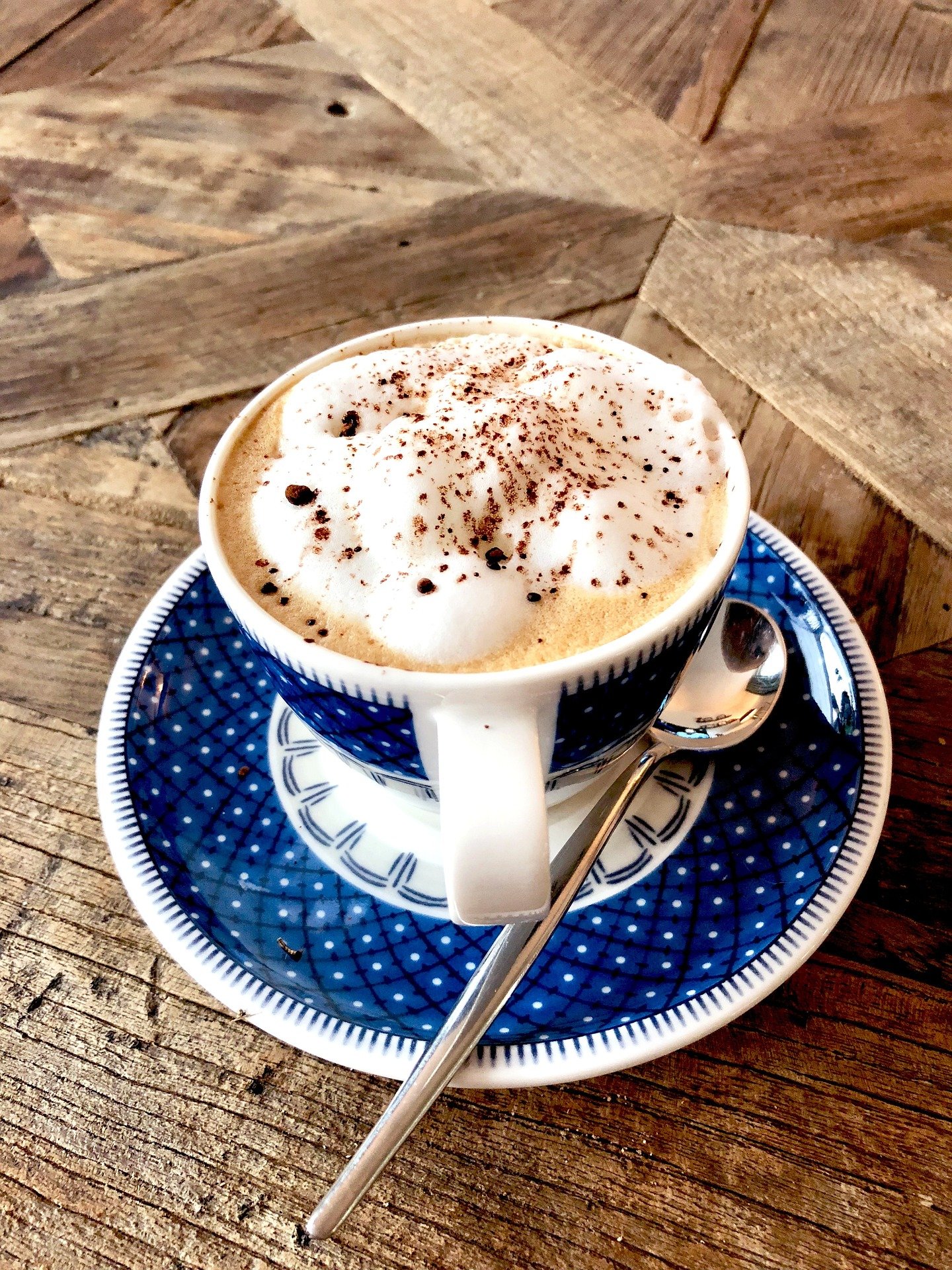A cup of coffee topped with whipped cream and cocoa powder, served on a blue patterned saucer with a silver spoon on a rustic wooden table.