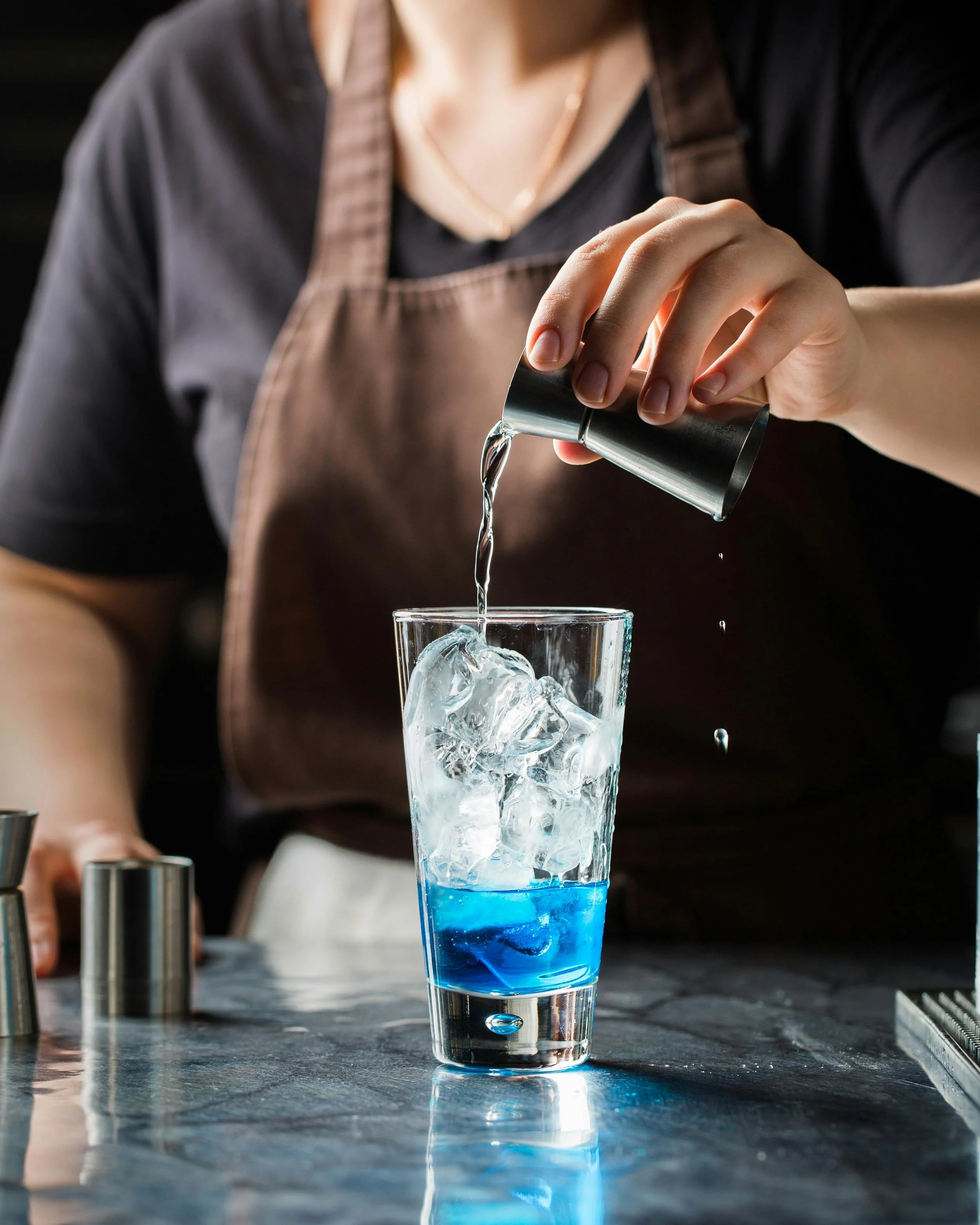 A person in a brown apron pours clear liquid into a tall glass filled with ice and a blue liquid at a bar or kitchen counter.