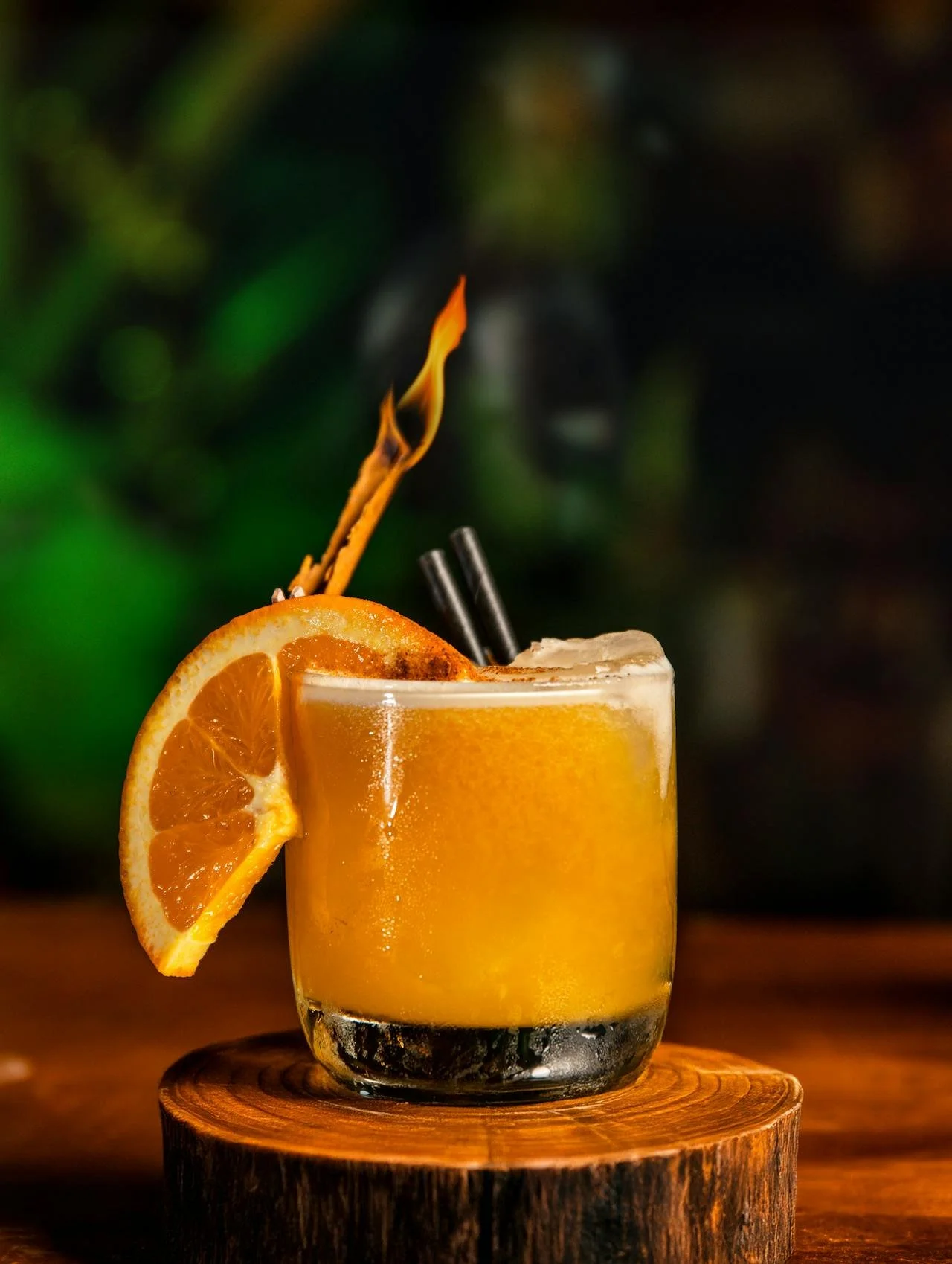 An orange cocktail garnished with a slice of orange, served in a short glass with a cinnamon stick and two small black straws, placed on a wooden coaster.