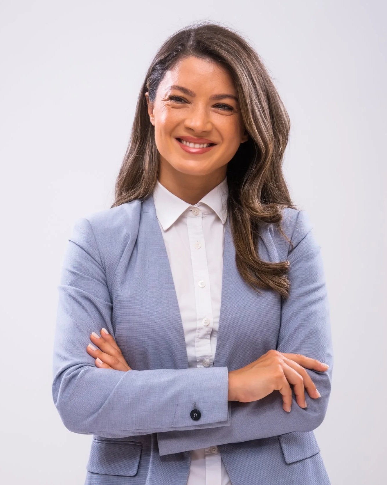 A smiling woman in a light grey blazer and white blouse with crossed arms against a plain background.