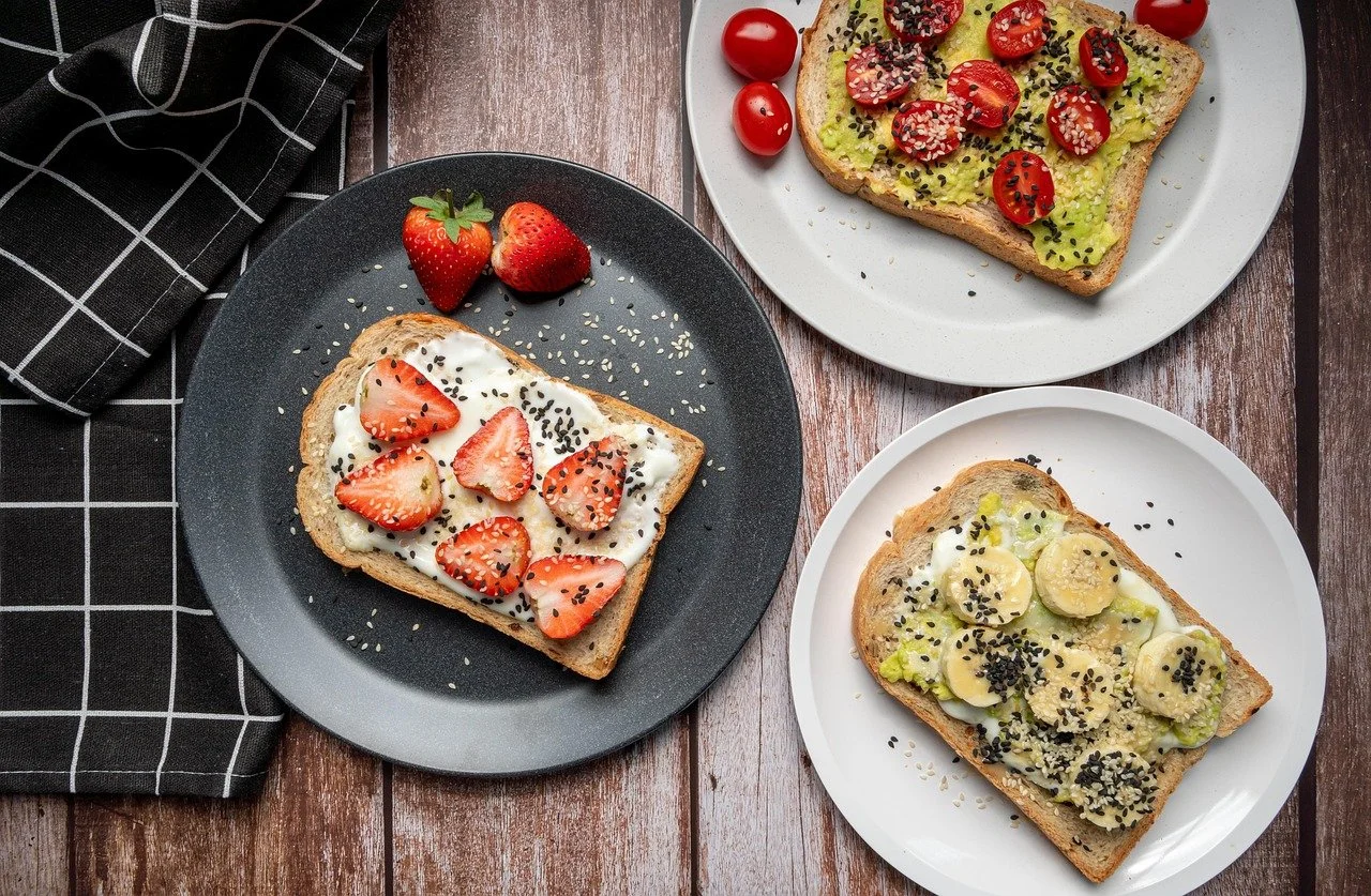 Three plates of toast with various toppings, including strawberries, cherry tomatoes, bananas, avocado, and black sesame seeds, on a wooden table with a black checkered napkin.