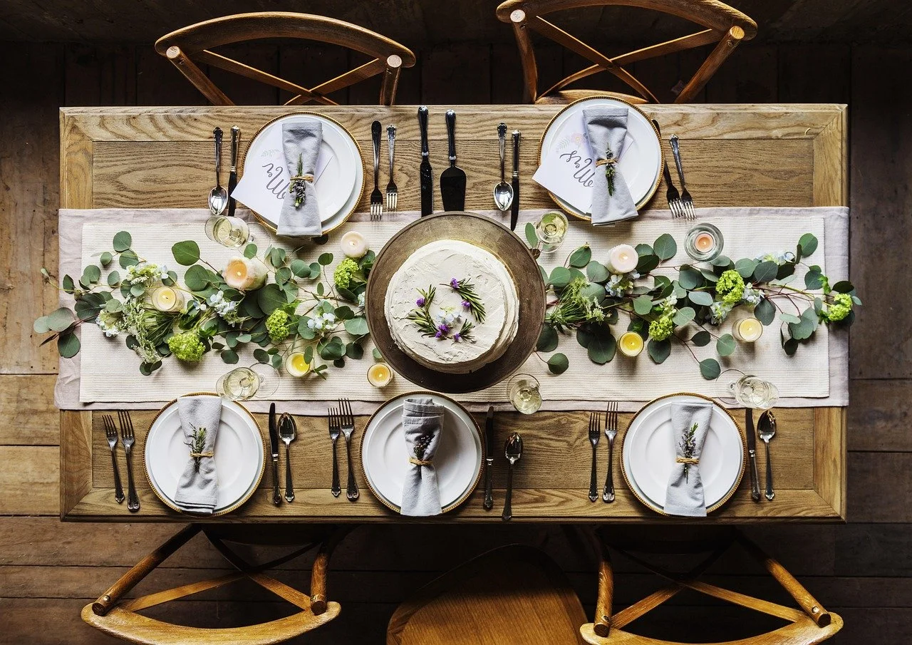 A top-down view of a dinner table setup with a cake in the center, surrounded by plates, cutlery, napkins, glasses, and floral decorations with candles.
