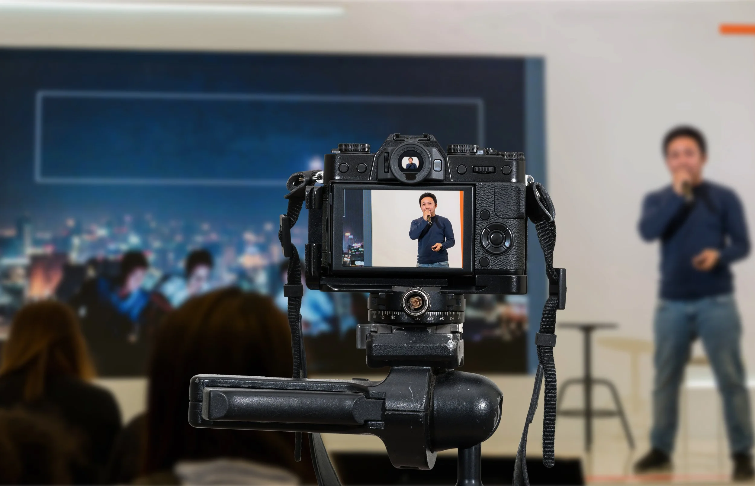 Camera recording a man speaking into a microphone during a presentation in a classroom or conference room.