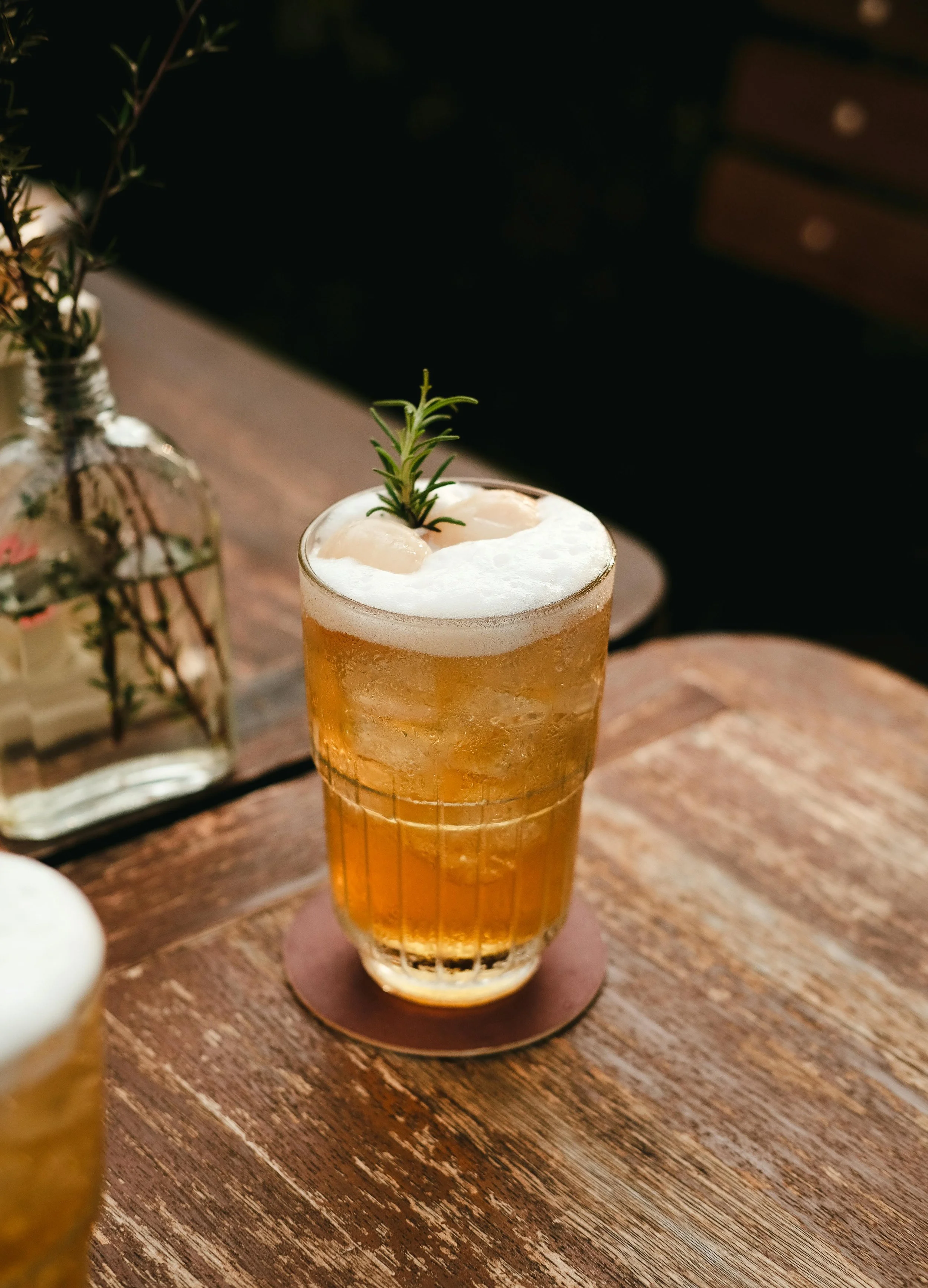A glass of beer with foam and a sprig of rosemary on top, on a wooden table with a vase of herbs in the background.