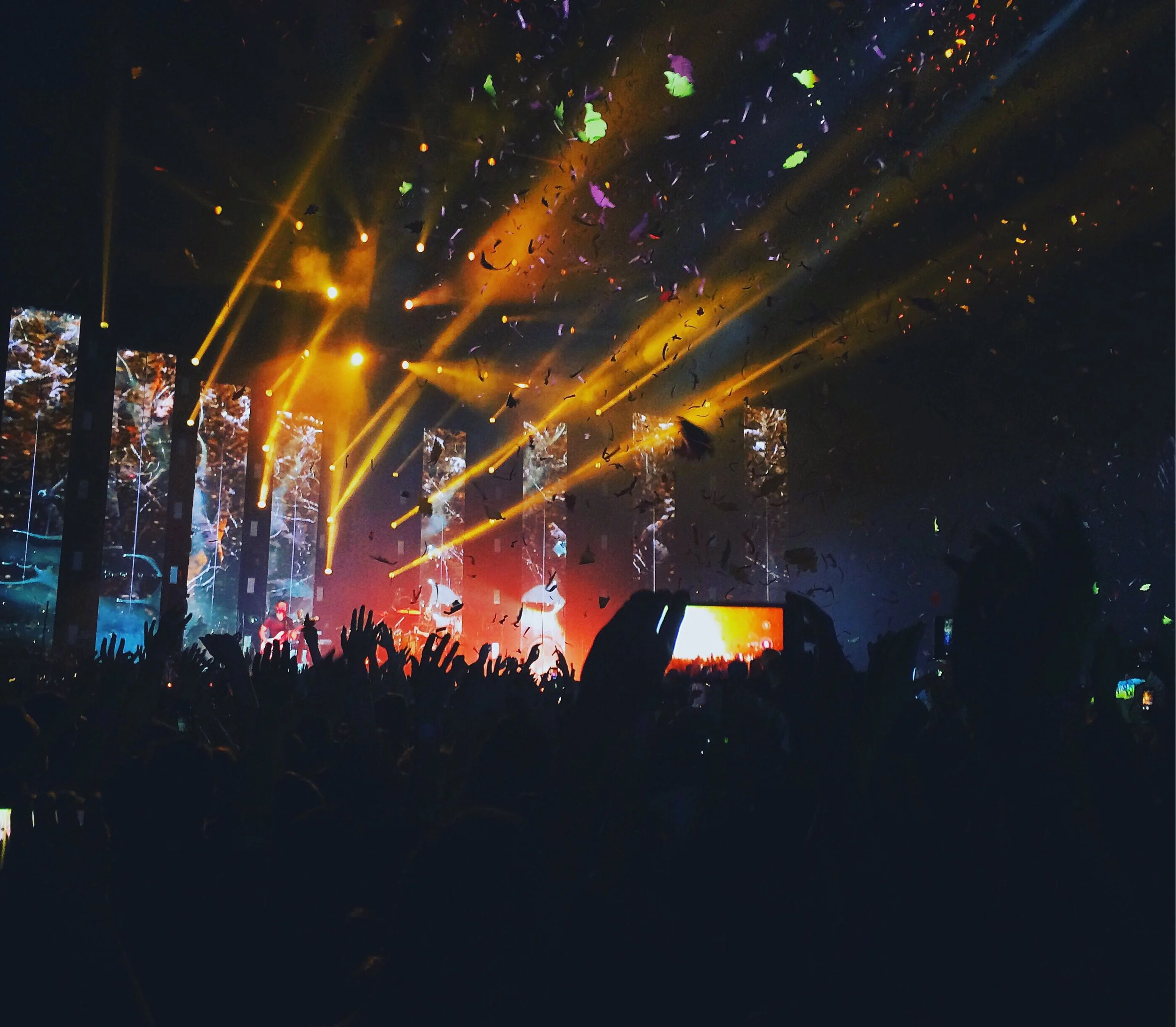 Concert scene with bright yellow lights, large digital screens showing visuals, and confetti falling over an audience with raised hands, capturing a lively performance.