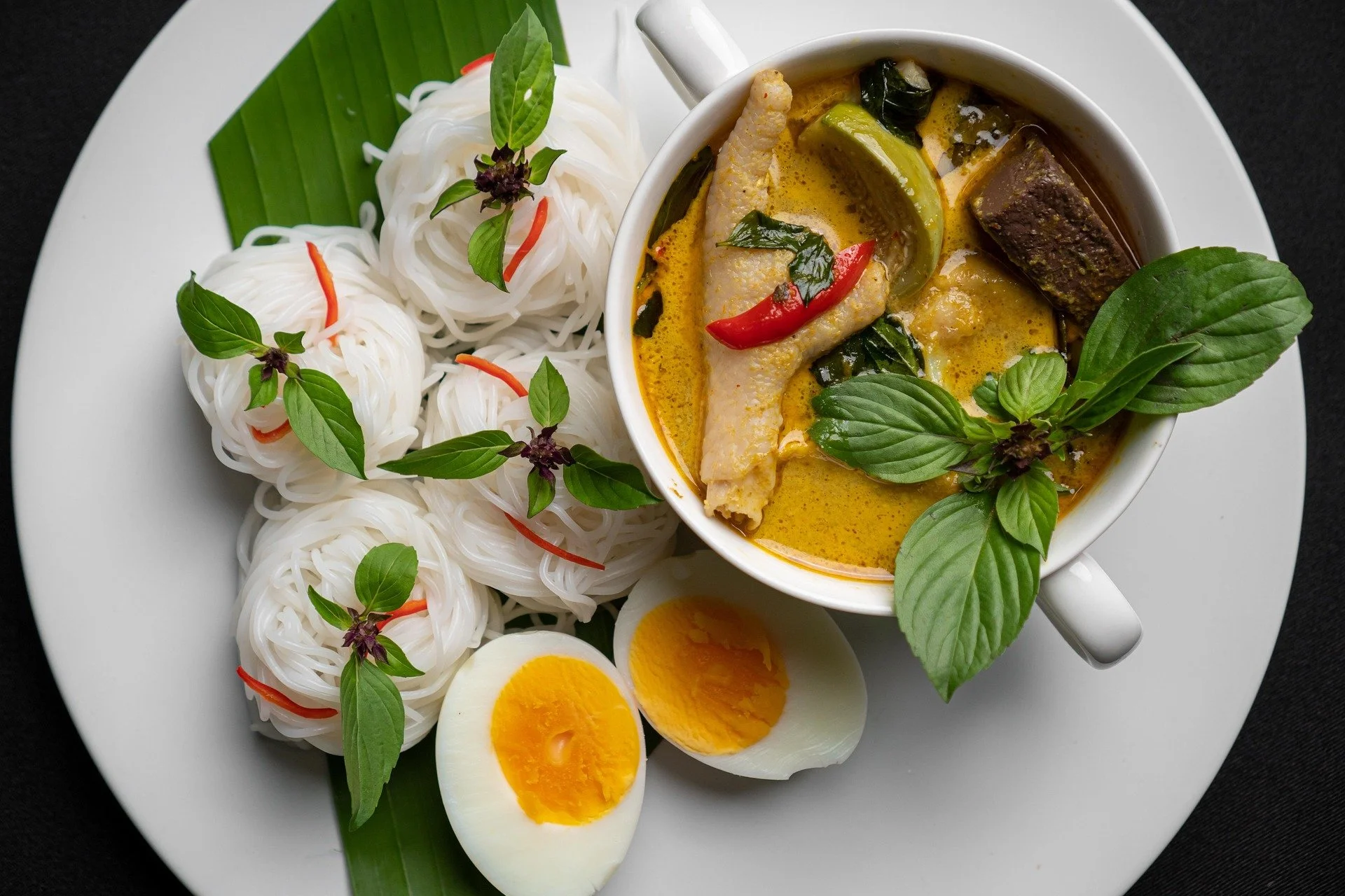Plate with rice noodles garnished with basil leaves, a cup of yellow curry with chicken, vegetables, chili peppers, and a halved boiled egg.