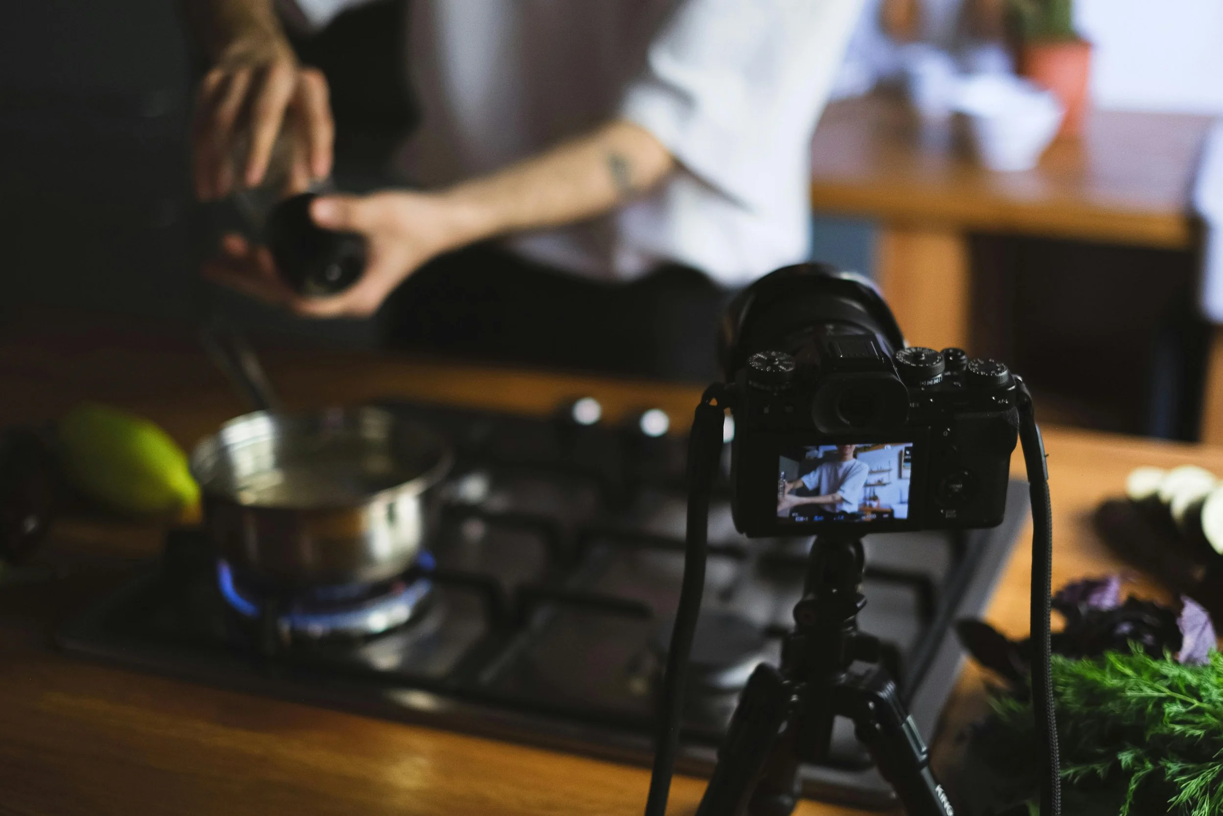 A person filming a cooking tutorial with a DSLR camera on a tripod in a kitchen, with ingredients and a stove on the counter.
