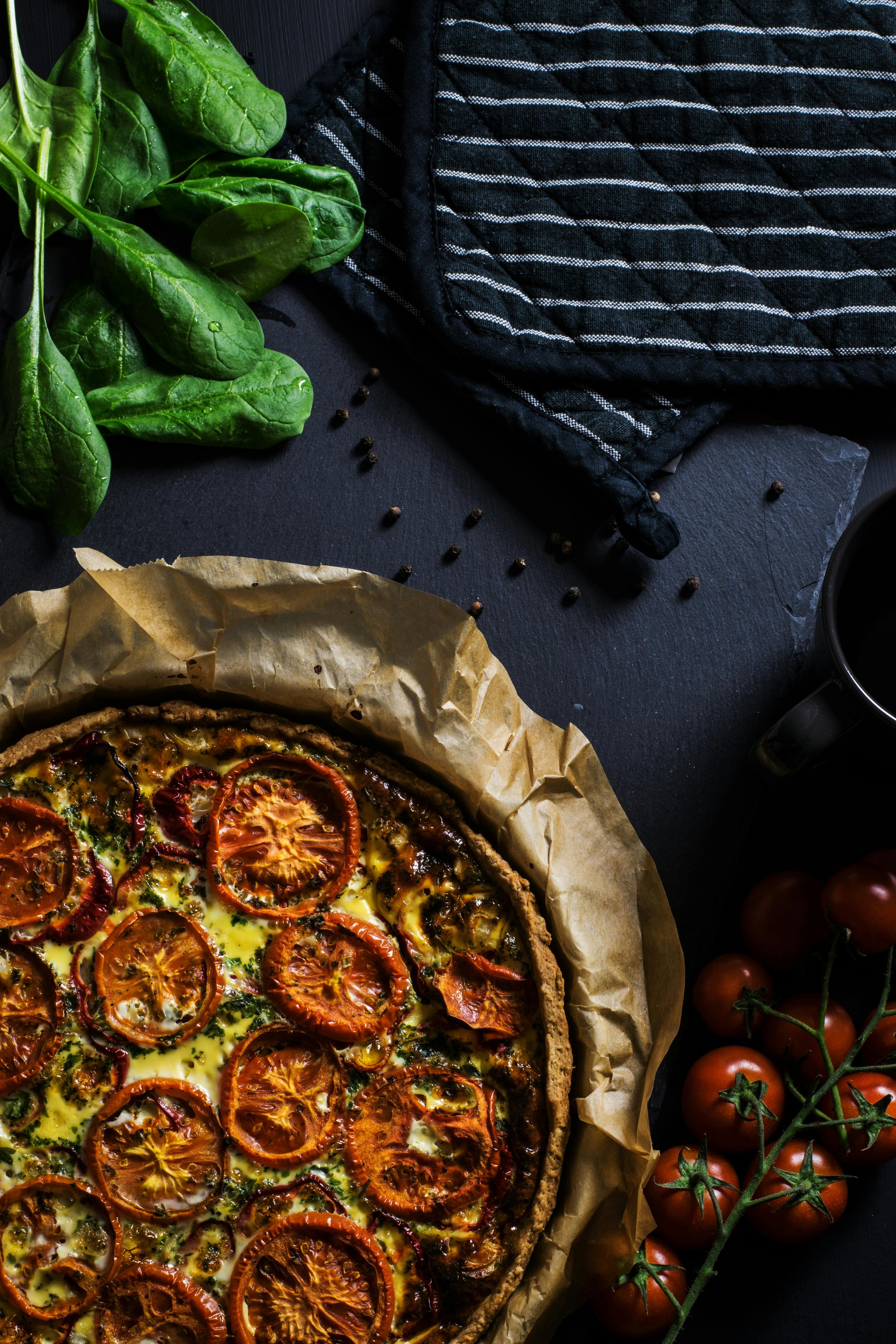 A freshly baked tomato tart on parchment paper, surrounded by cherry tomatoes on the vine, fresh spinach, a black mug, and a striped oven mitt on a dark surface.