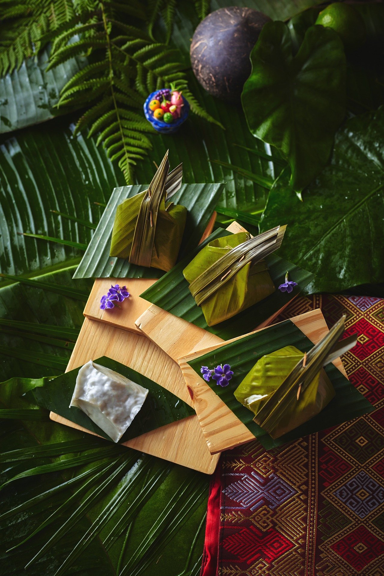 Traditional Southeast Asian Festive Offerings with banana leaf packaging, purple flowers, wooden tray, surrounded by lush tropical greenery and textured cloth.