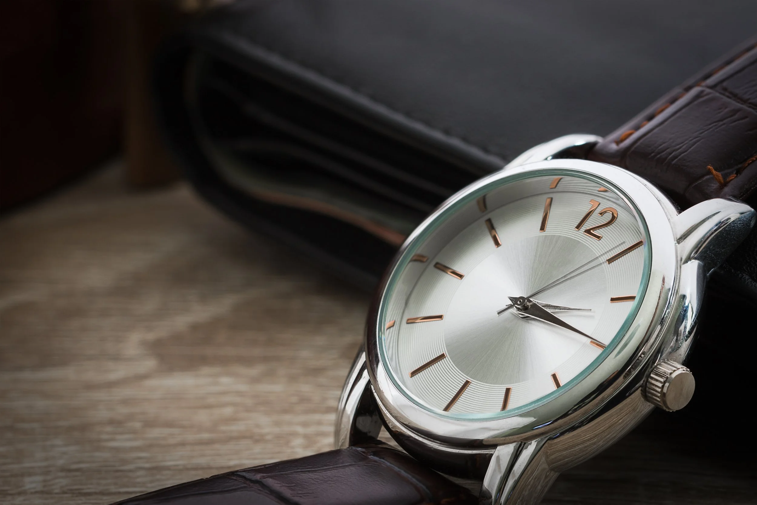 Close-up of a silver wristwatch with a brown leather strap, showing the time as approximately 4:23, placed on a wooden surface with a black leather wallet in the background.