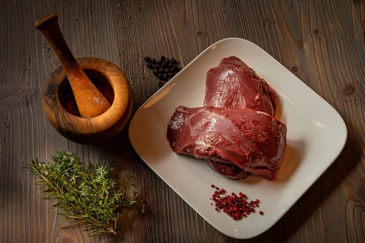 Two raw beef steaks on a white plate, a sprig of thyme, a small pile of peppercorns, and a wooden mortar with a pestle on a wooden surface.