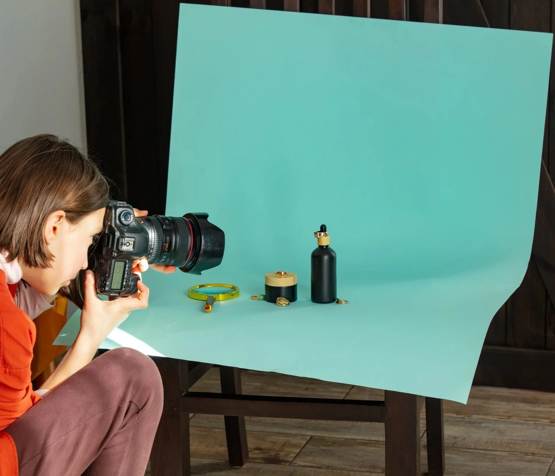 A woman with short brown hair holding a professional camera, taking pictures of jewelry displayed on a blue background, including a black bottle with gold accents, a black and gold container, and earrings.