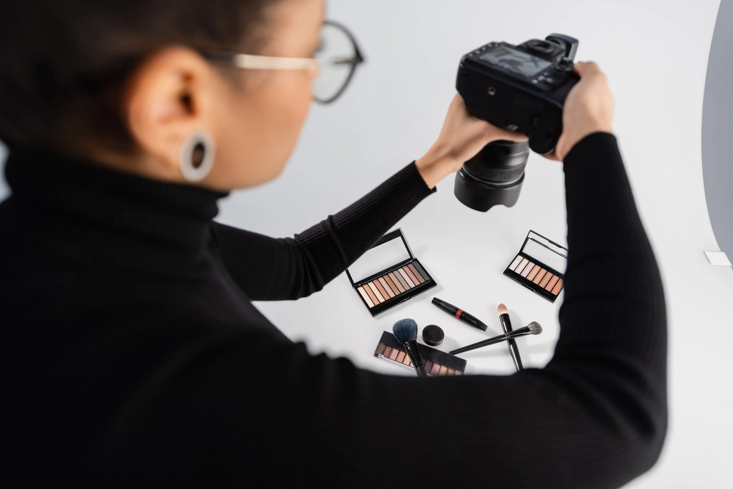 A person takes a photo of makeup products, including eyeshadow palettes, brushes, and lip products, arranged on a white surface.