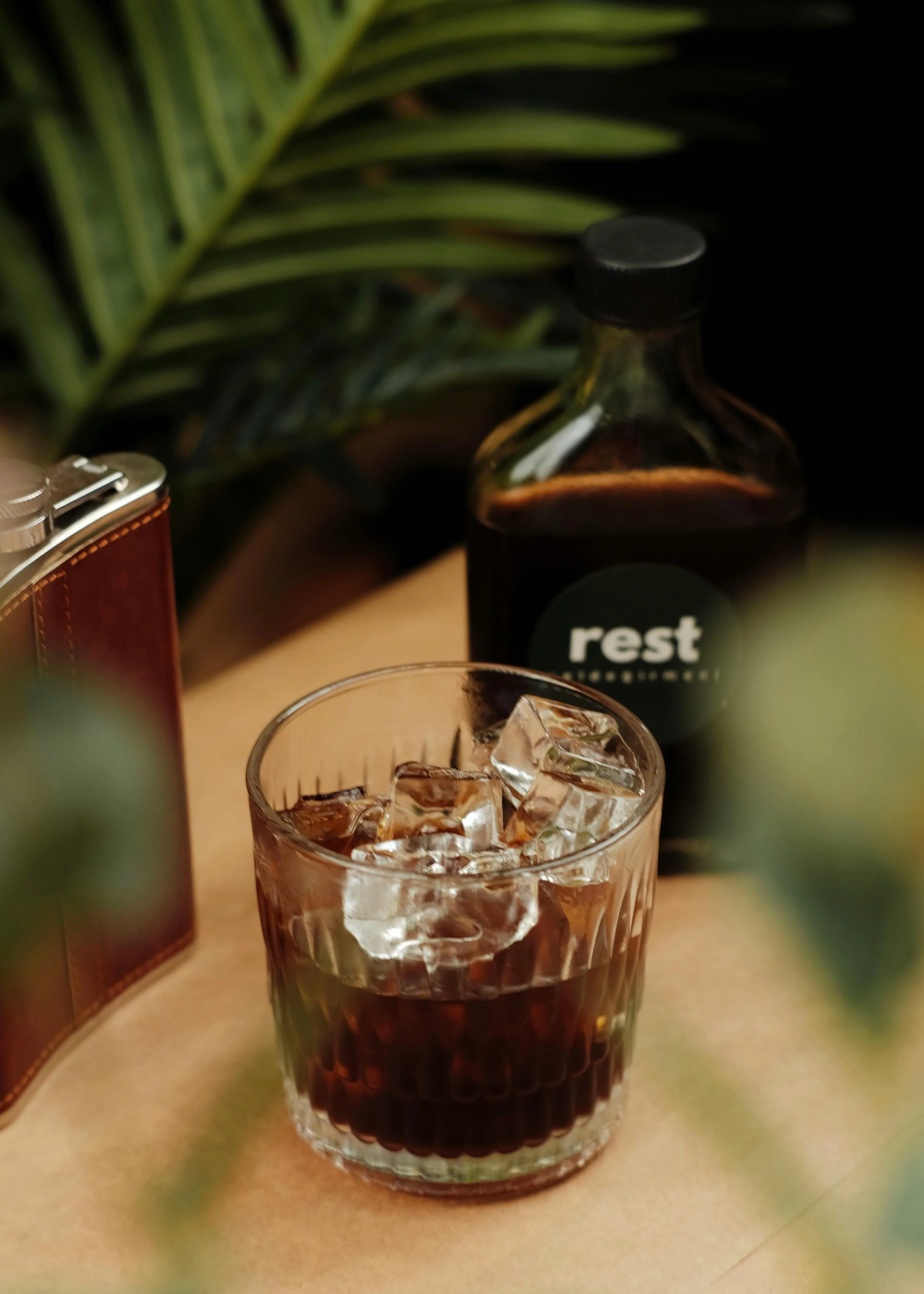 A glass of dark soda with ice cubes, a bottle labeled 'rest,' a vintage leather wallet, and a blurred green plant in the background.
