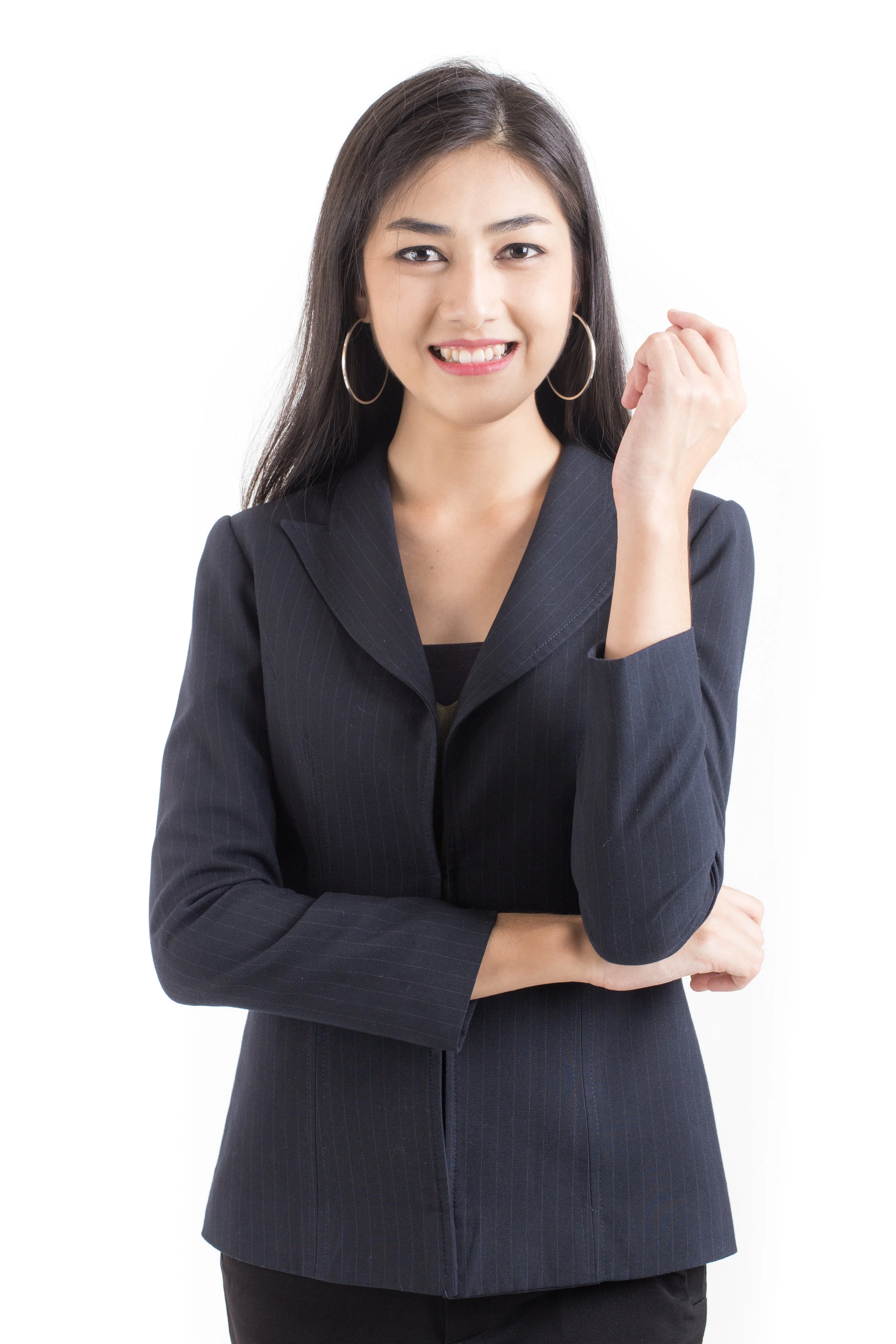 A young woman with long dark hair, wearing a black blazer and hoop earrings, smiling and making a fist gesture against a white background.