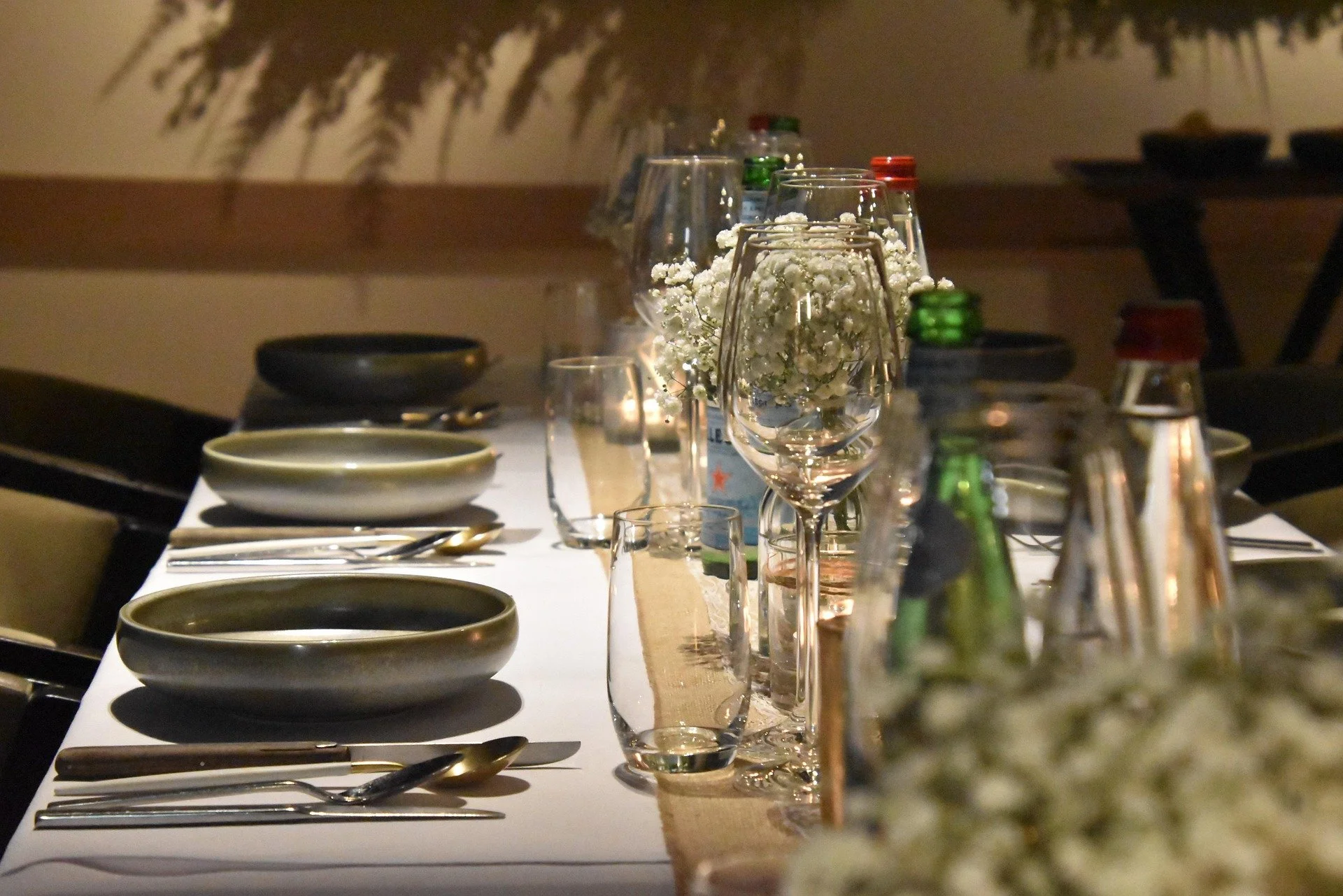 A elegantly set dining table with black bowls, gold-colored utensils, crystal glasses, and small floral centerpieces, with a beige table runner in a dimly lit room.