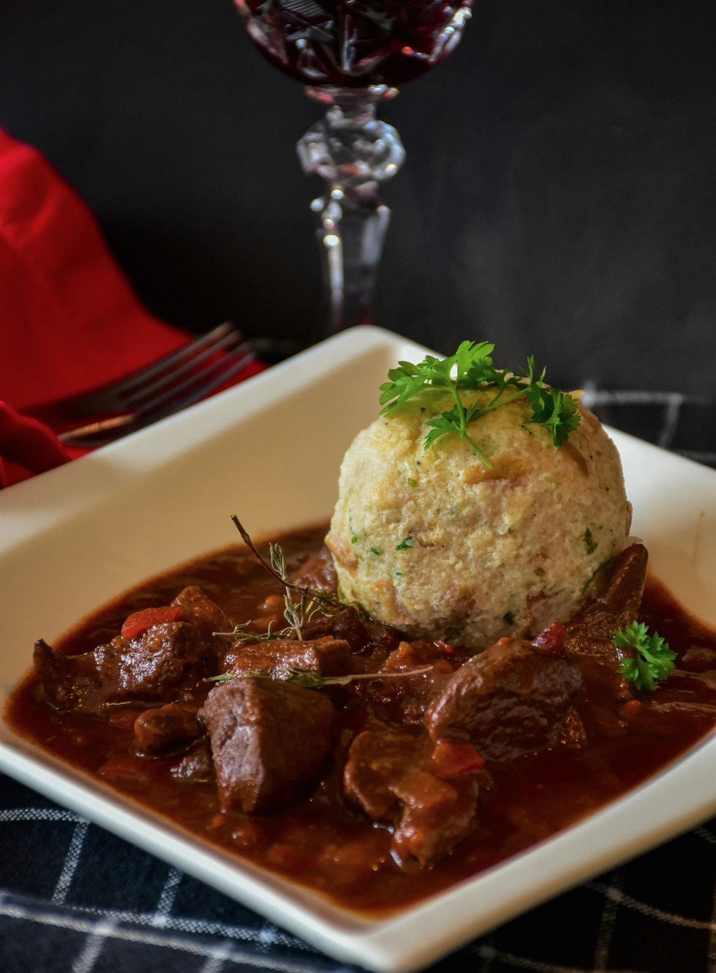 A square white plate with beef stew and a large scoop of mashed potatoes garnished with parsley, set on a black tablecloth with a wine glass in the background.