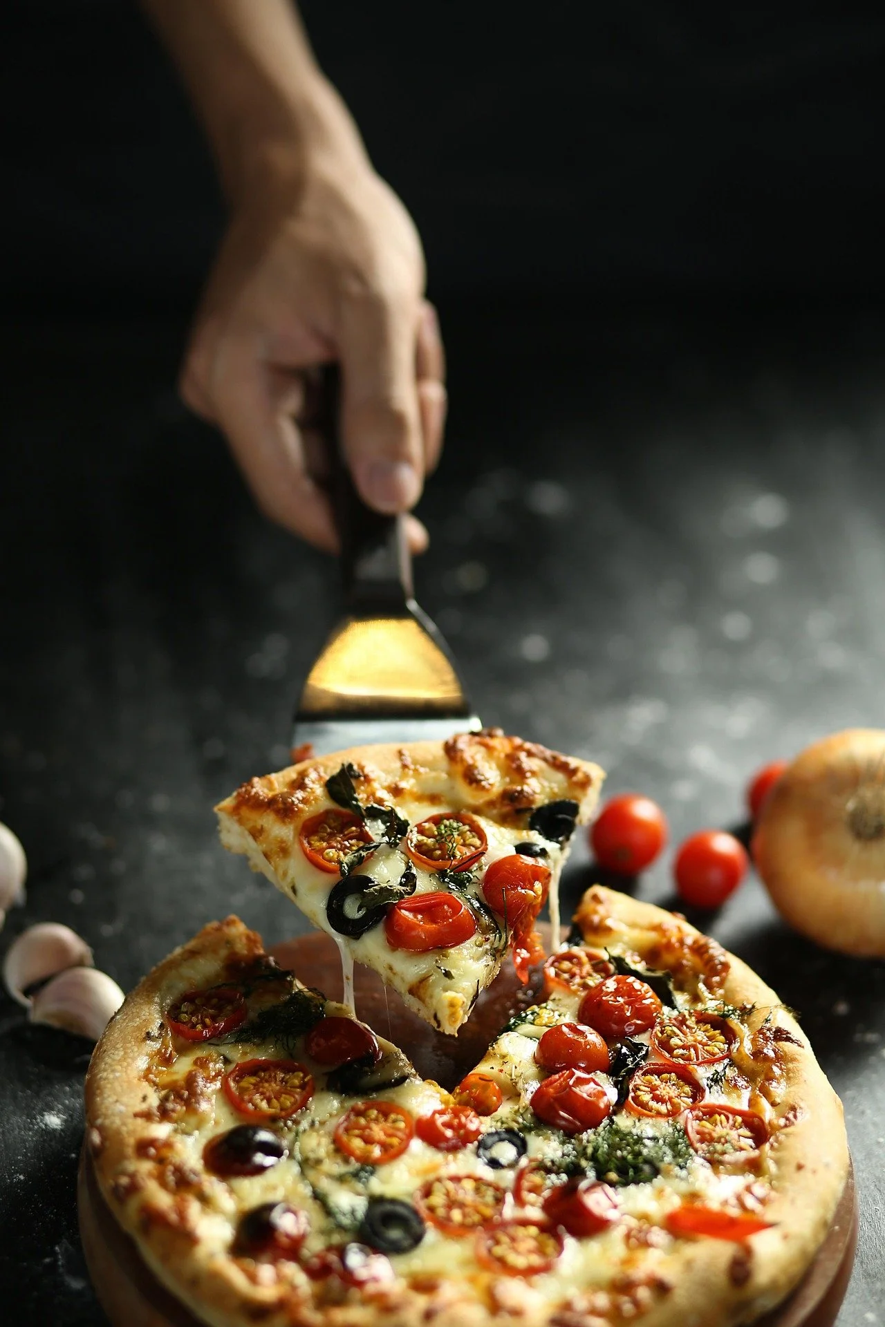A hand is holding a pizza cutter and slicing a veggie pizza topped with cherry tomatoes, black olives, and herbs. The pizza sits on a wooden serving board on a dark surface with garlic cloves, cherry tomatoes, and an onion nearby.