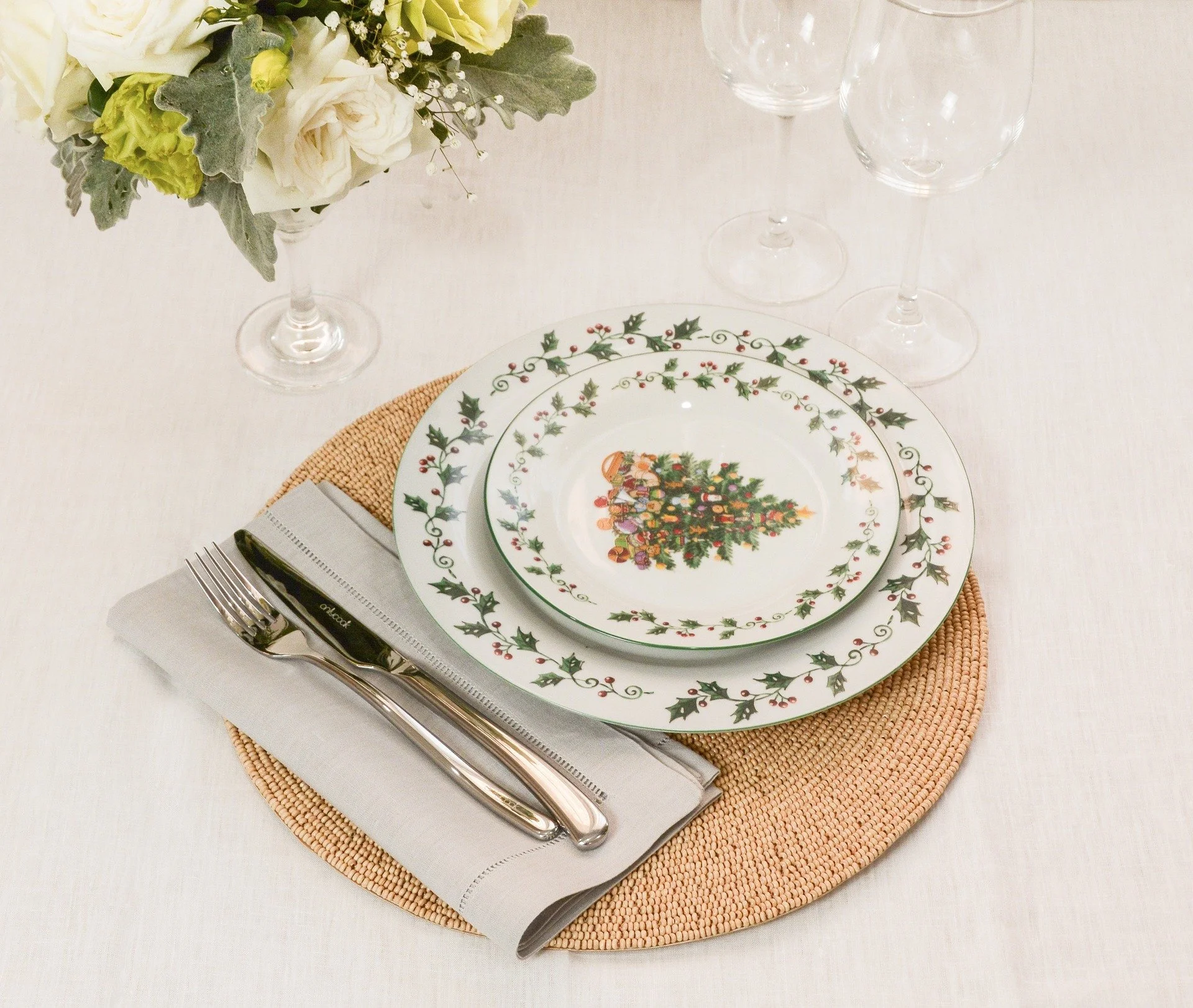 A festive holiday table setting with Christmas-themed dinnerware, a wedding or banquet centerpiece, a woven placemat, a folded napkin, and silverware, with empty wine glasses and a floral arrangement with white and green flowers on a white tablecloth