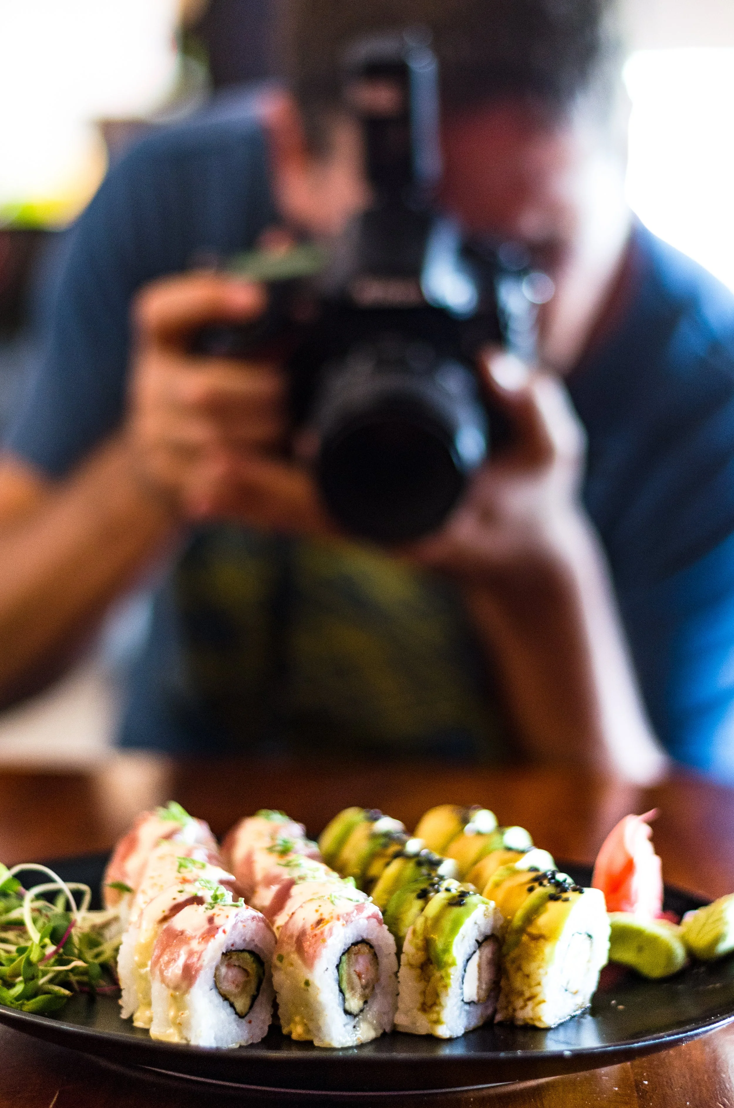 A person taking a photo of a plate of sushi rolls with a professional camera, sitting at a table in a restaurant.