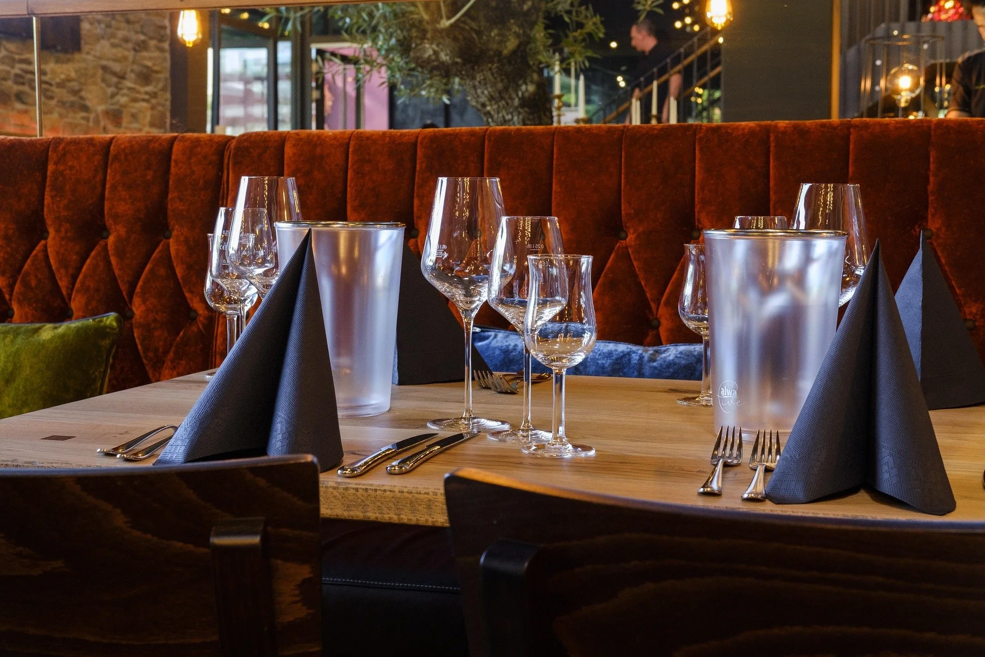 A restaurant table set with wine glasses, water glasses, black napkins, silverware, and frosted water pitchers, with a plush red velvet booth in the background.