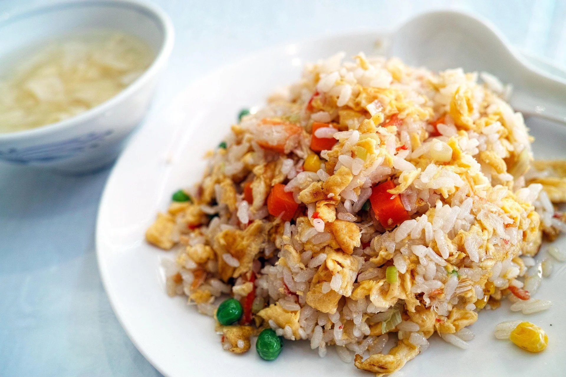 Close-up of a plate of fried rice with vegetables and scrambled eggs, accompanied by a bowl of clear soup in the background.