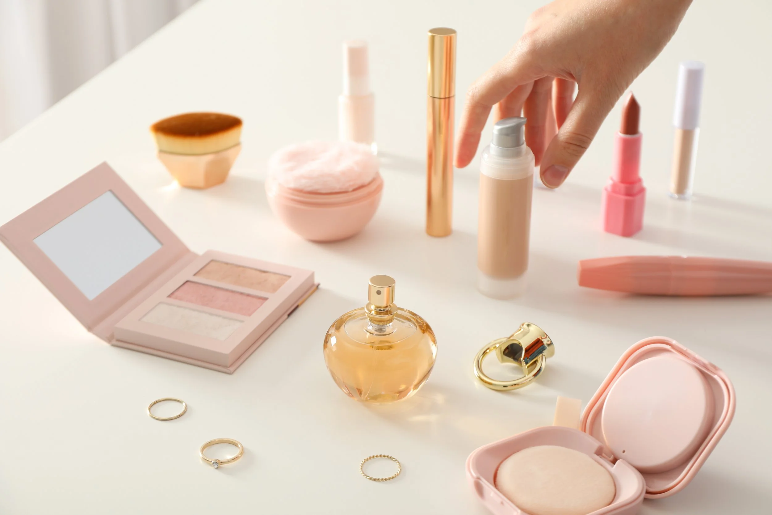 A collection of makeup and jewelry items on a white surface, including blush, perfume, rings, lip balm, and various beauty products.