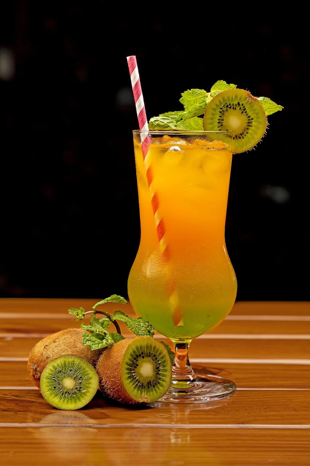 A tall glass of orange and yellow layered fruit smoothie garnished with a slice of kiwi, fresh mint leaves, and a red and white striped straw, placed on a wooden table with whole and sliced kiwis beside it.