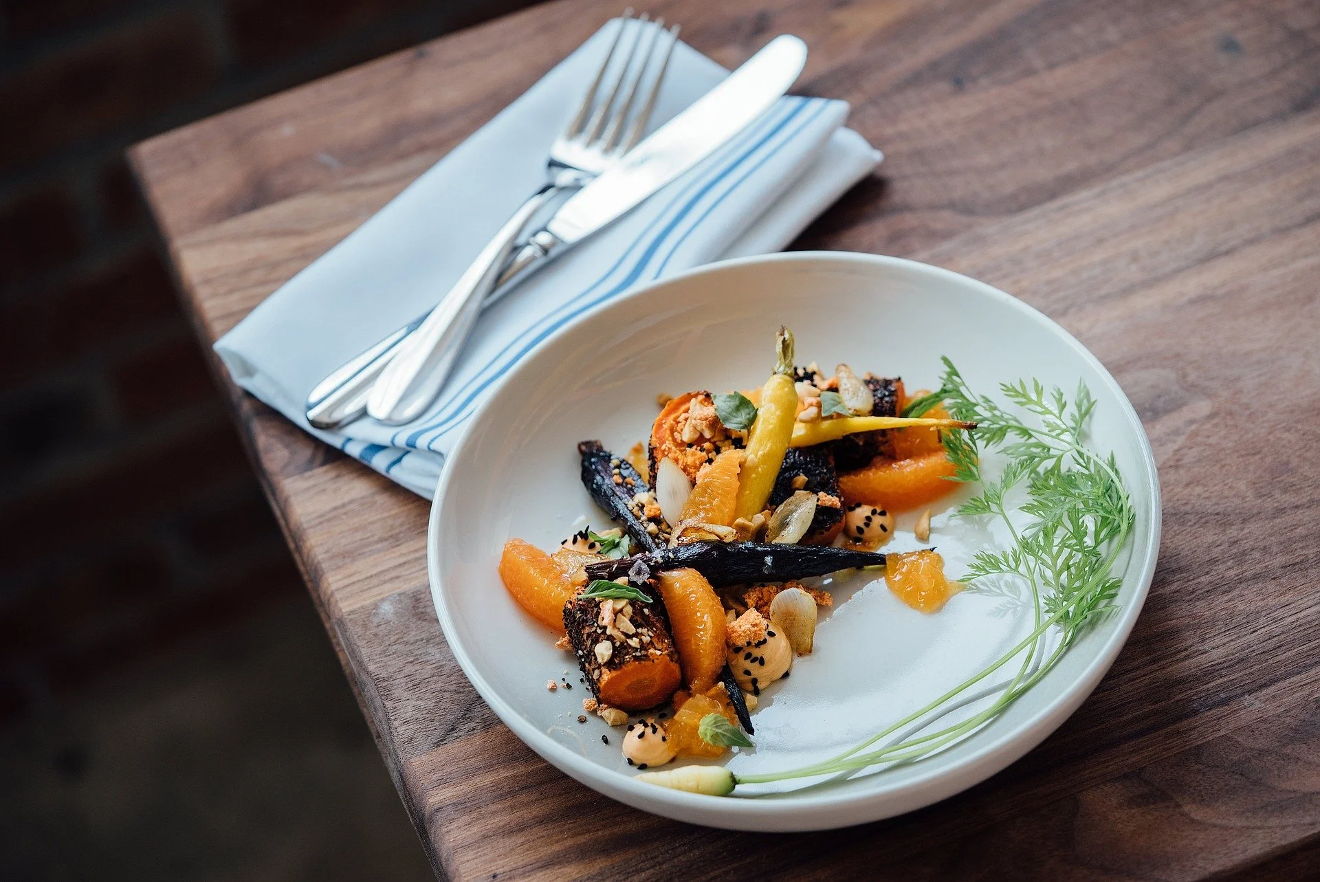 A white plate with roasted vegetables, including carrots, yellow peppers, and garnished with greens, served with a sprinkle of nuts and seeds, on a wooden table beside a wrapped set of silverware with a blue-and-white napkin.