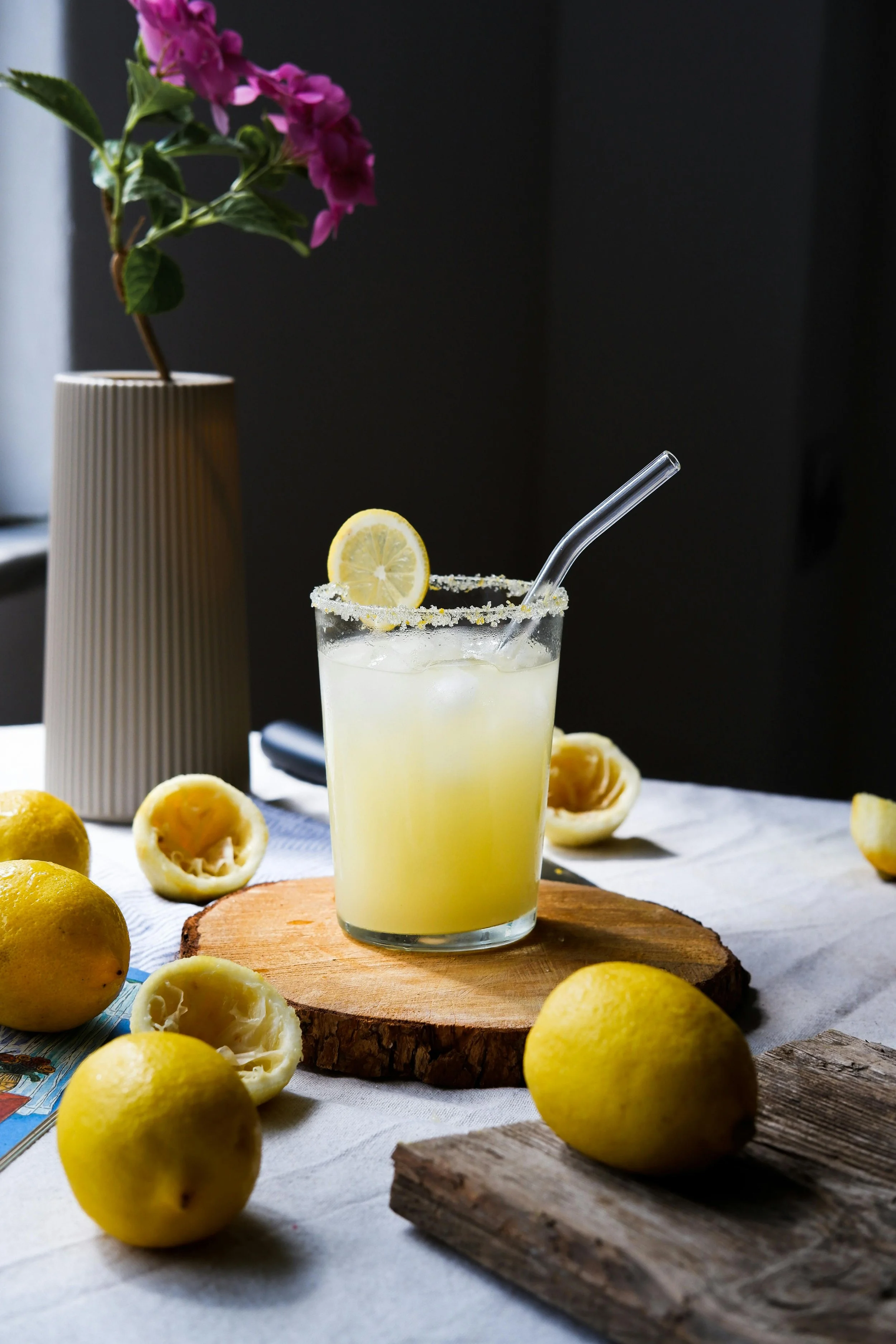 A glass of lemonade with a lemon slice and a straw, surrounded by whole and cut lemons on a rustic table, with a potted plant with pink flowers in the background.