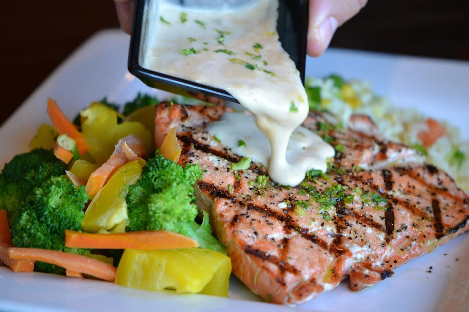 Salmon fillet being drizzled with white creamy sauce onto a bed of grilled vegetables including broccoli, carrots, and yellow squash on a white plate.