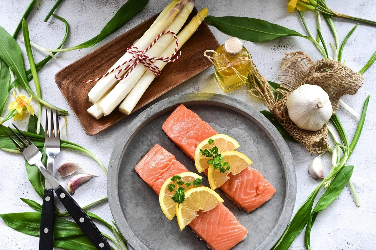 Raw salmon fillets garnished with lemon slices and herbs on a gray plate, surrounded by garlic, asparagus, oil, and green leaves.