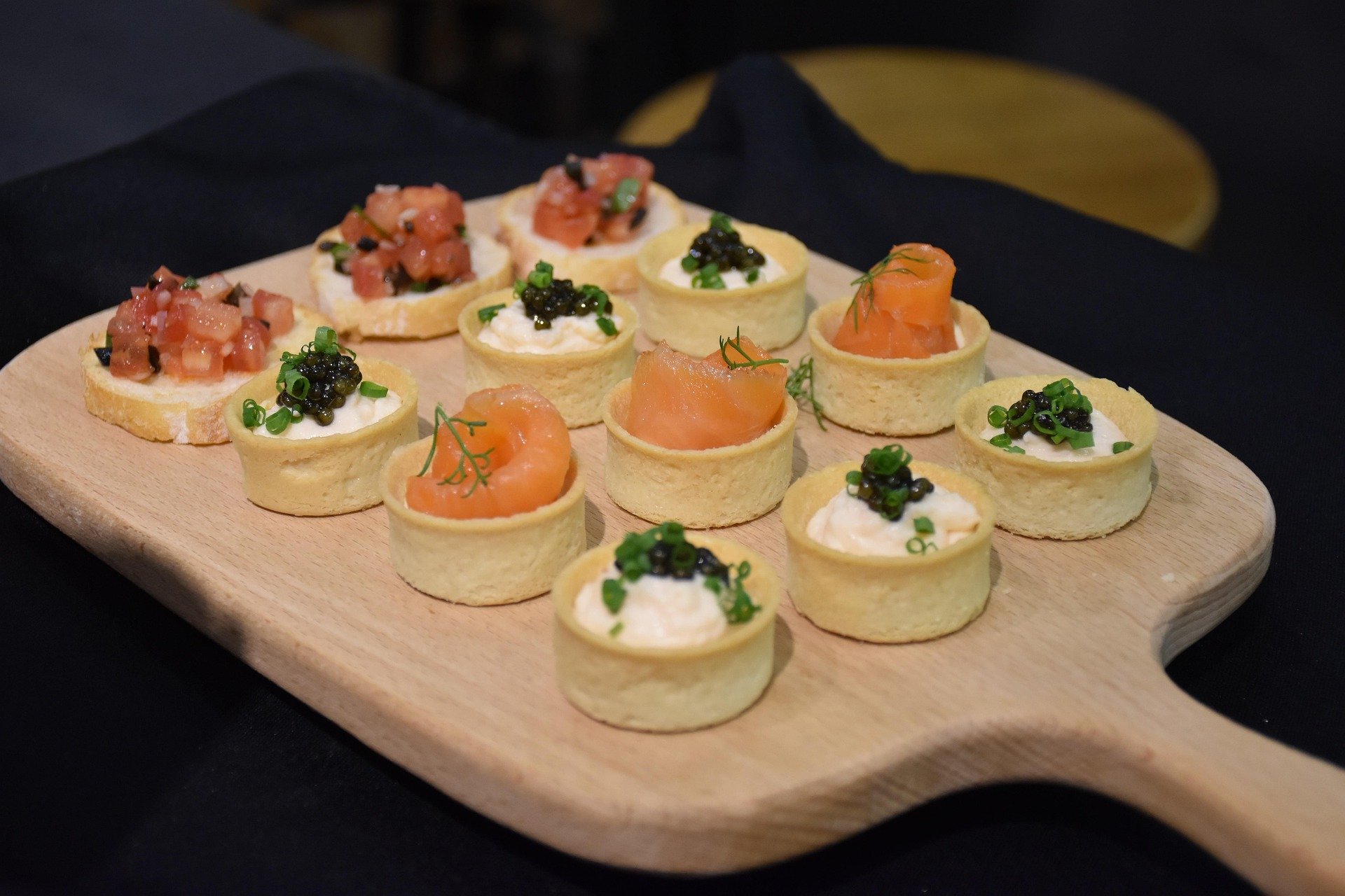 Assorted bite-sized appetizers on a wooden serving board, including small tart shells filled with cream cheese and topped with smoked salmon and herbs, and crispy pastry bases topped with diced raw fish, green onions, and caviar.