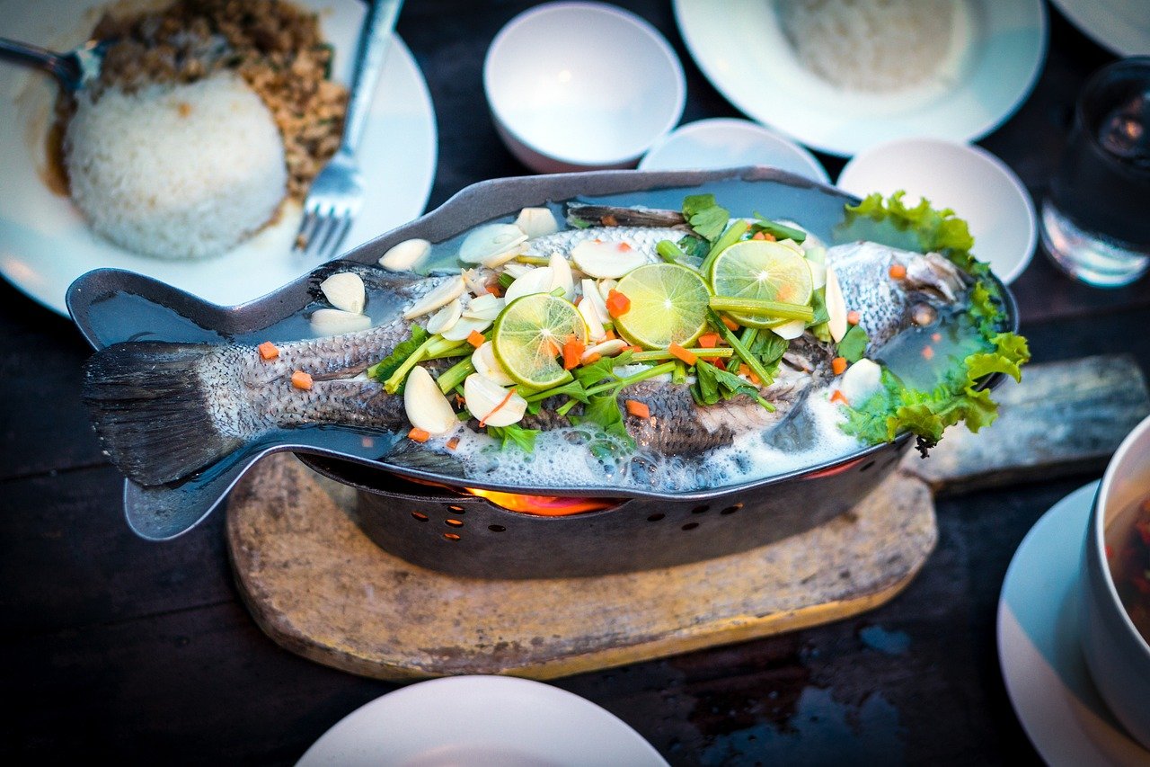 Whole fish garnished with lime slices, garlic, herbs, and vegetables, served on a black platter with a side of rice and other dishes around it.