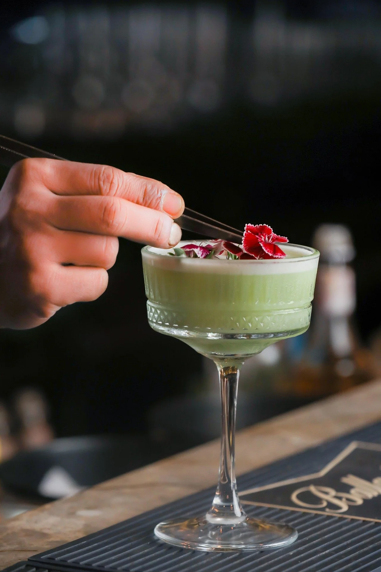 A bartender garnishes a green cocktail in a coupe glass with a red flower.