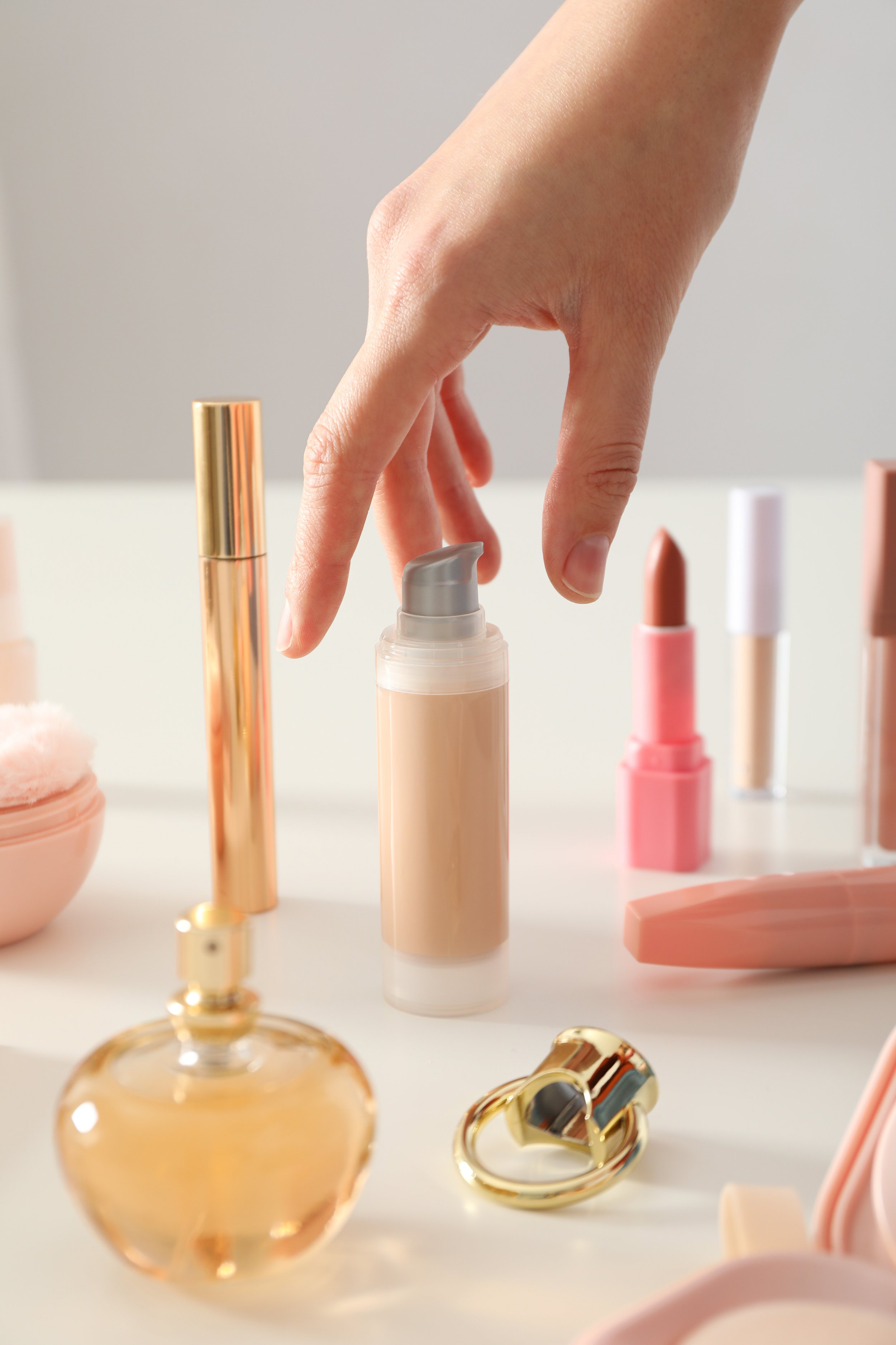 A hand reaching for a beige pump bottle of facial moisturizer or sunscreen on a white surface surrounded by makeup and beauty products, including lipstick, a perfume bottle, and a compact mirror.