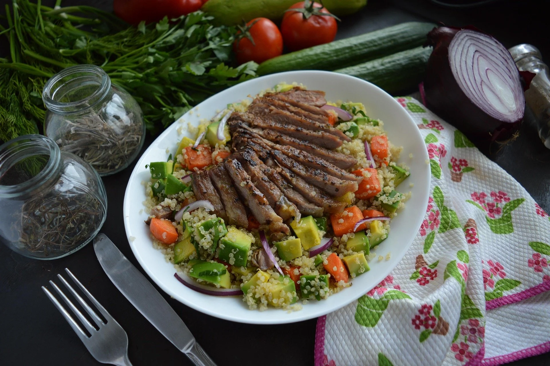 A plate of quinoa salad topped with sliced grilled meat, surrounded by chopped vegetables, on a black table with fresh produce and herbs in the background.