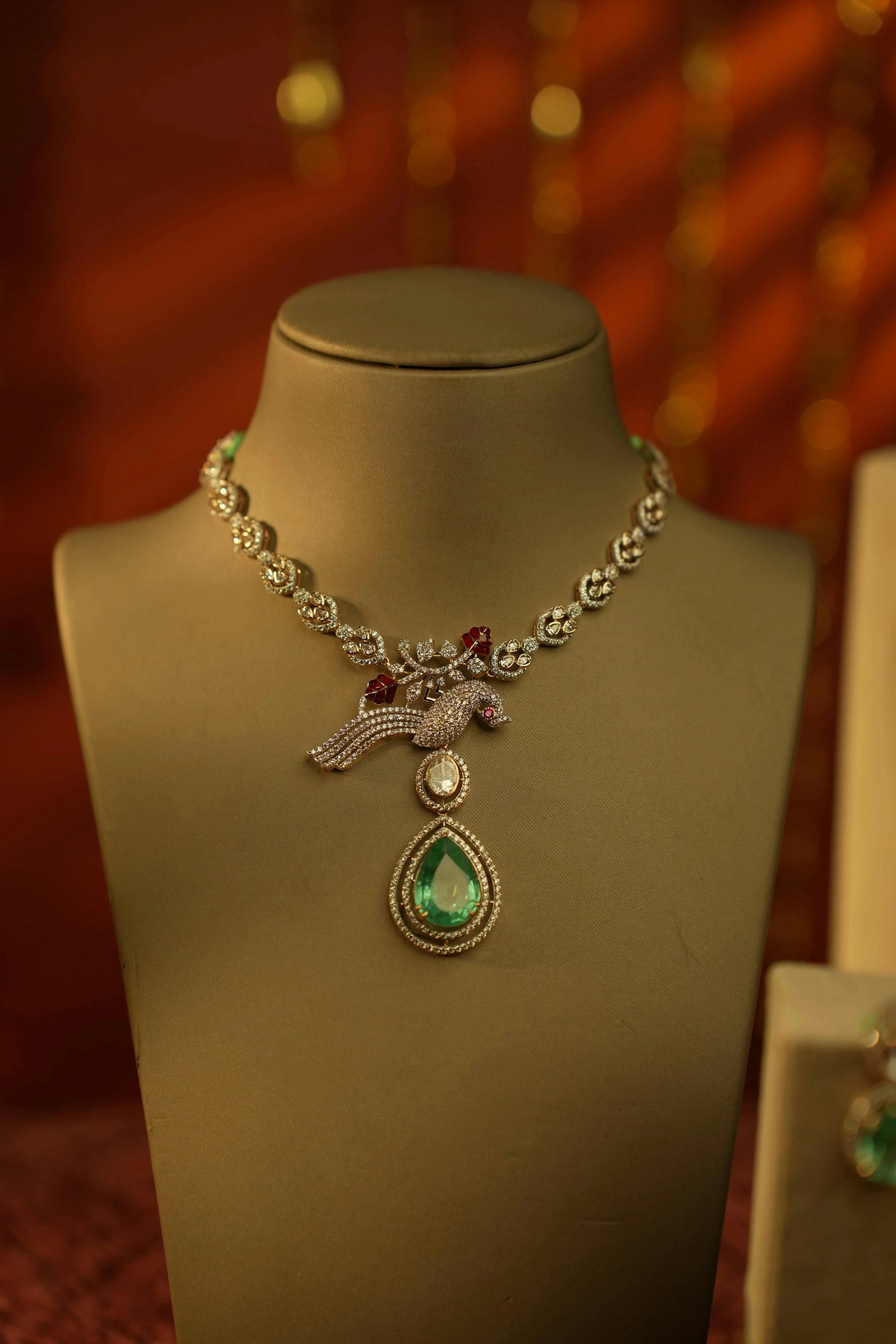 Jewelry display with a diamond necklace featuring a bird motif and large green gemstone pendant, set against a blurred warm background.