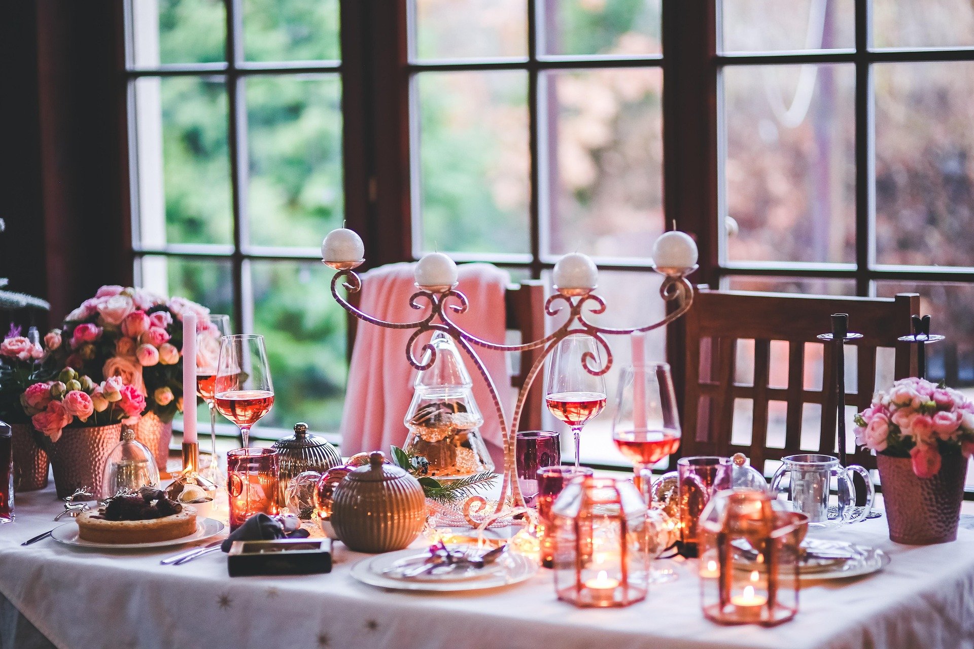 A dining table decorated with pink flowers, candles, wine glasses with rosé wine, and a candelabra, set near large windows with a view of greenery outside.