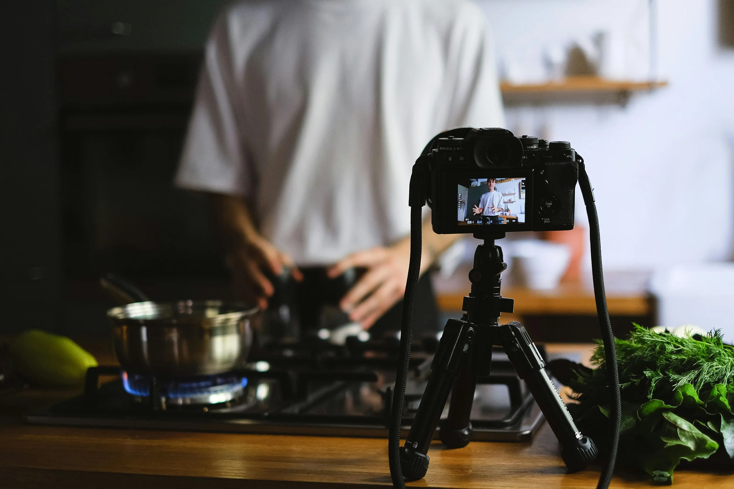 A camera on a tripod recording a person cooking in a kitchen, with fresh greens on the counter.