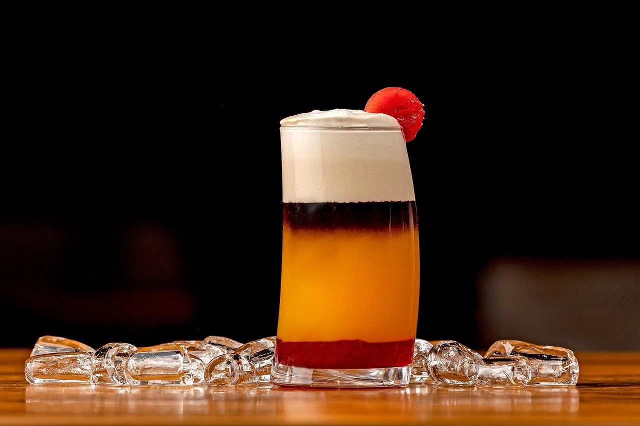 Colorful layered cocktail with foam and a cherry garnish on top, placed on a wooden surface with ice cubes around it, against a black background.